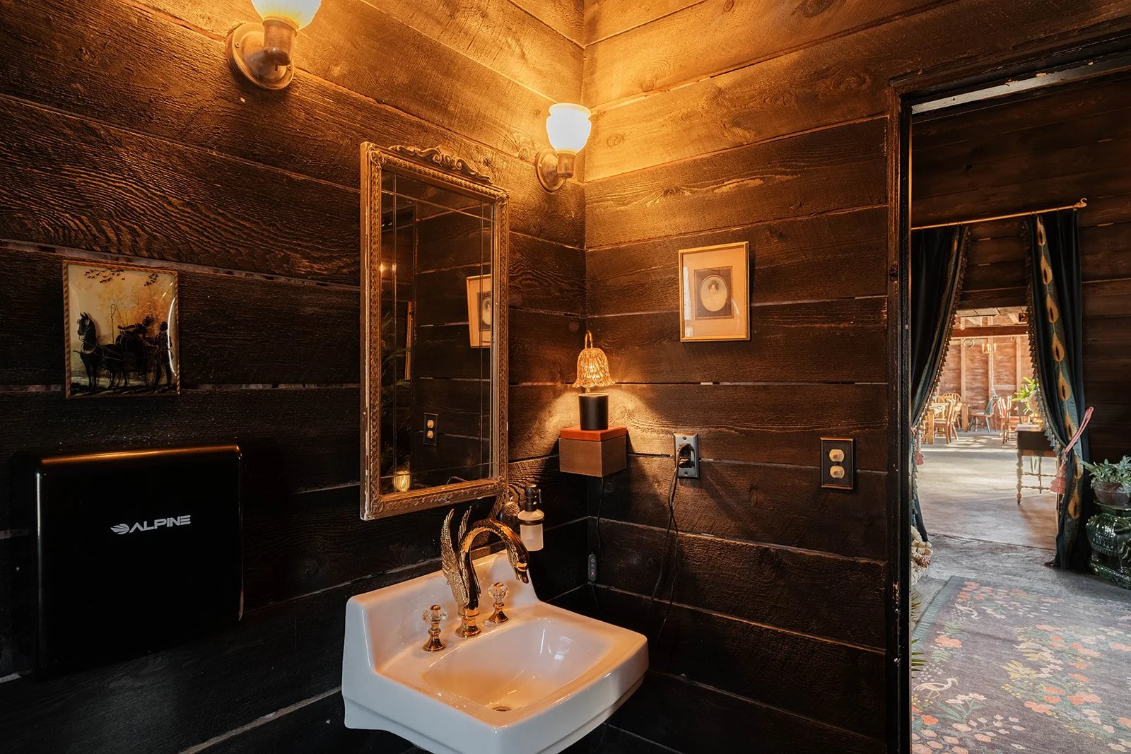 A small rustic bathroom with dark wooden walls, a white sink with gold fixtures, a mirror, a small framed picture, a wall-mounted lamp, and an adjacent room with floral curtains and outdoor seating visible through an open doorway.