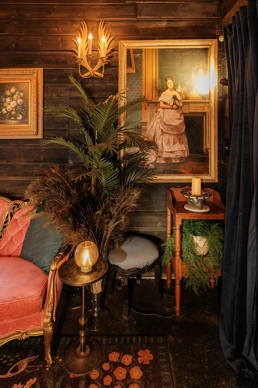 Vintage-style interior with a pink tufted sofa, a small lamp, a plant, a black stool, a wooden table with green fern, candles, ornate framed paintings on wood-paneled wall, and dark curtains.