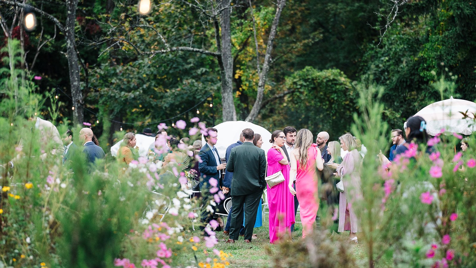 People attending an outdoor garden party or wedding reception, gathered in groups, with string lights overhead, surrounded by greenery and flowers.