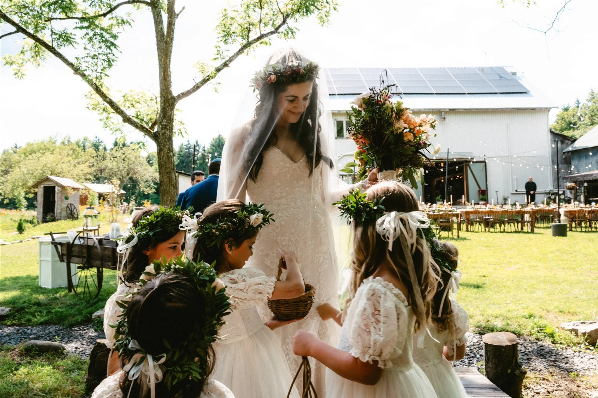 A bride in a lace dress and flower crown surrounded by flower girls on the lawn in back of the barn at Black Walnut Farm, a Catskills Wedding venue