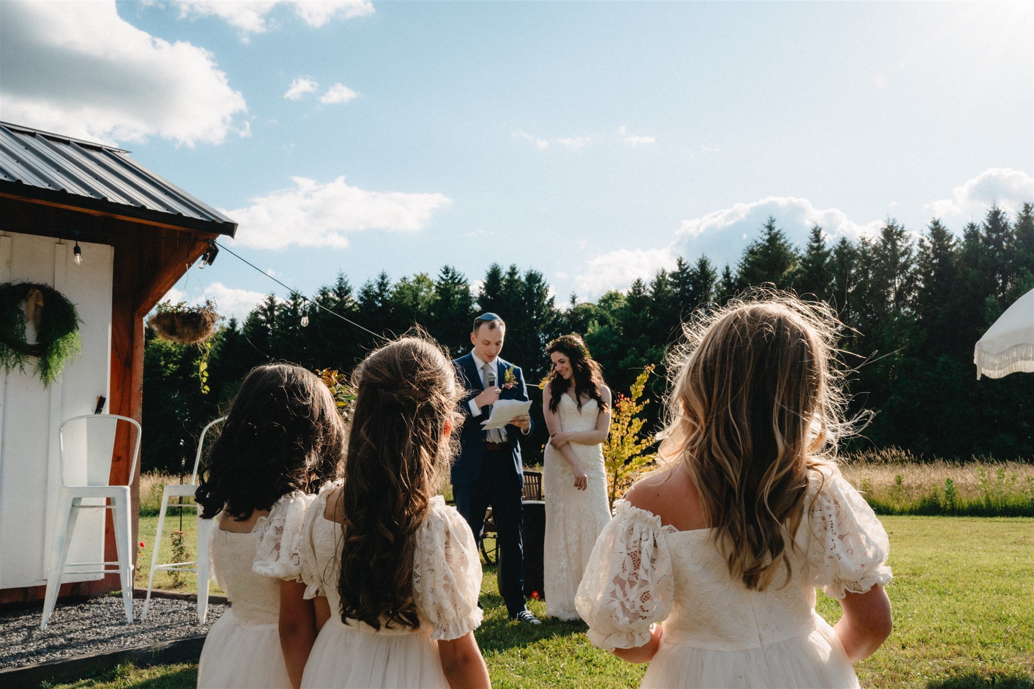 A wedding ceremony outdoors with six women, four young girls in white dresses, and two adults, a man and a woman, in formal attire, standing in front of a wooden building, in a grassy field with trees and a blue sky with clouds.