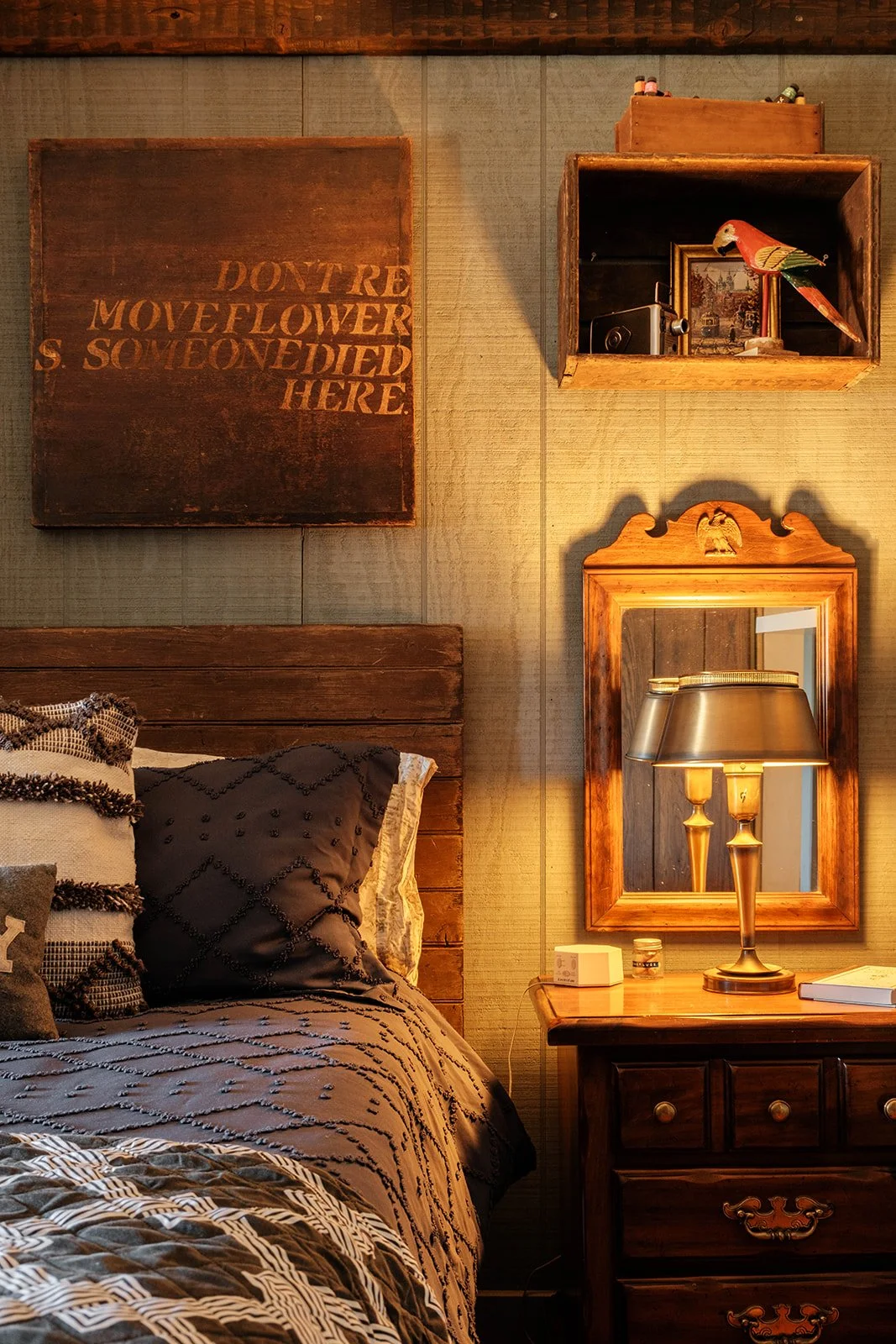 A cozy bedroom corner featuring a wooden headboard, a nightstand with a lamp, a mirror, and framed artwork and decorative items on the wall.