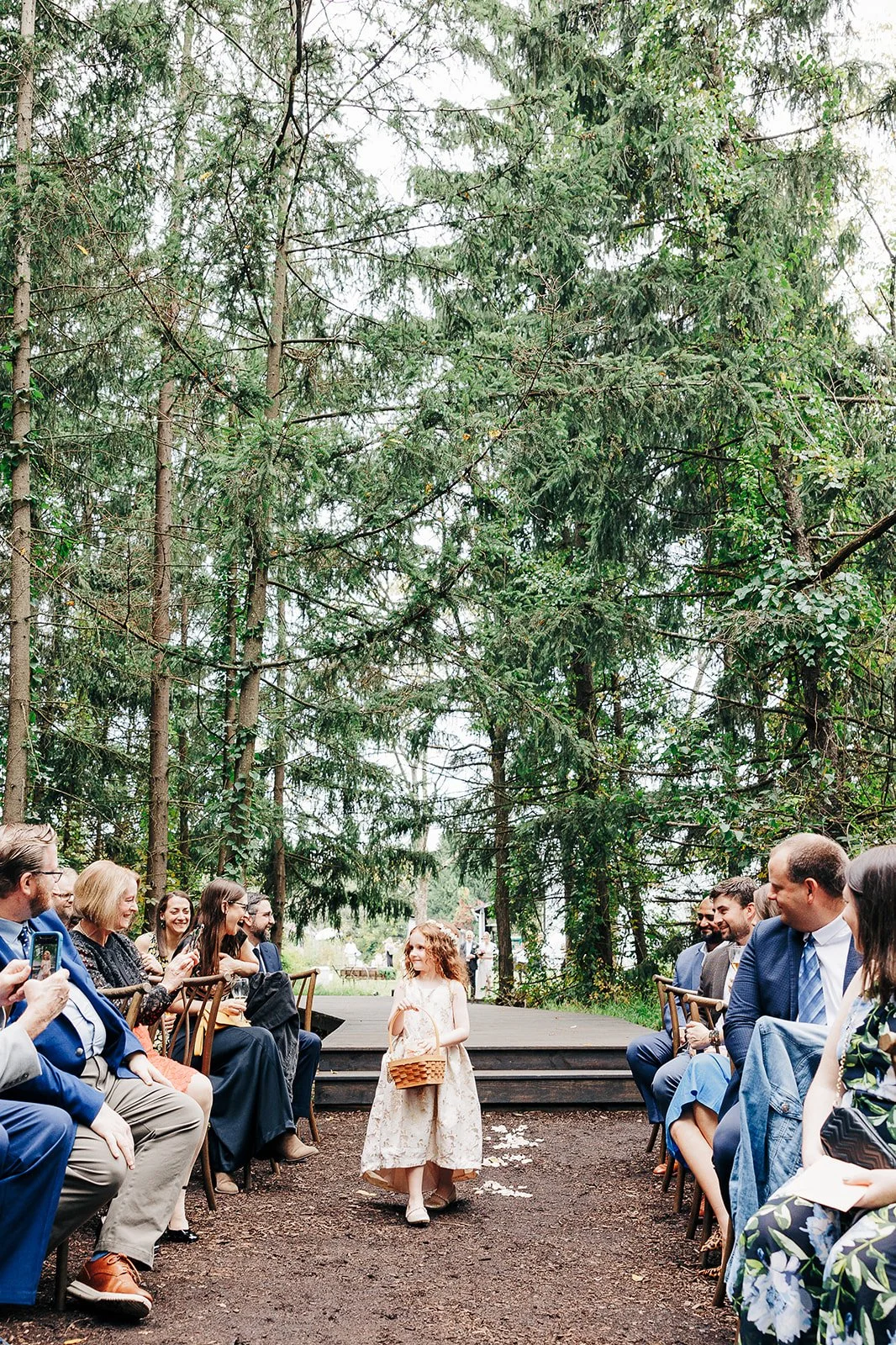 A young girl in a floral dress walking down an outdoor aisle holding a basket, surrounded by seated guests at a wedding ceremony in a forest setting.