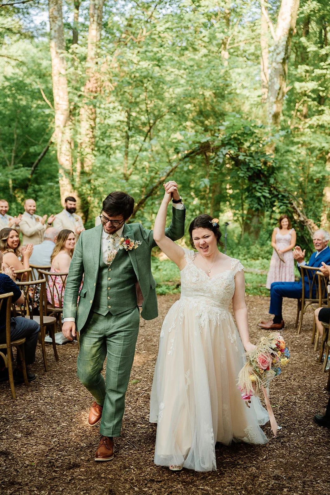 A wedding couple walking arm-in-arm in a forest clearing, with the bride holding a bouquet, as guests seated on either side applaud and smile.