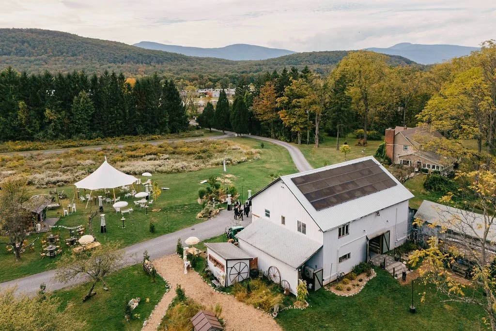 A rural outdoor setting with a white barn, decorated with outdoor furniture and a white canopy tent, surrounded by trees and hills in the background, during fall.