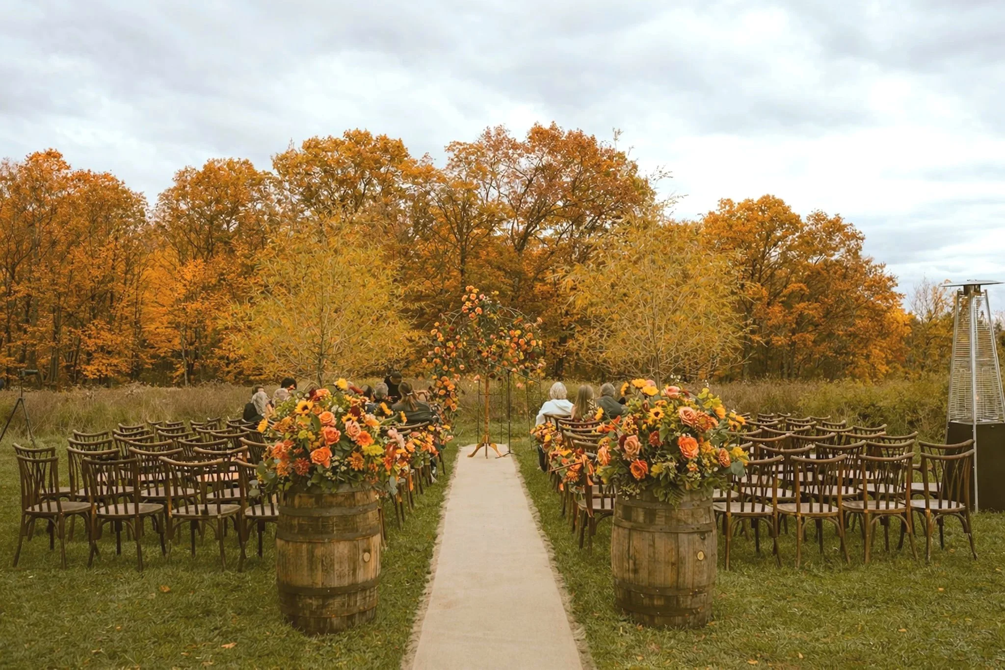 Outdoor wedding ceremony setup on a grassy field with rows of chairs, large floral arrangements in barrels, and autumn trees in the background.