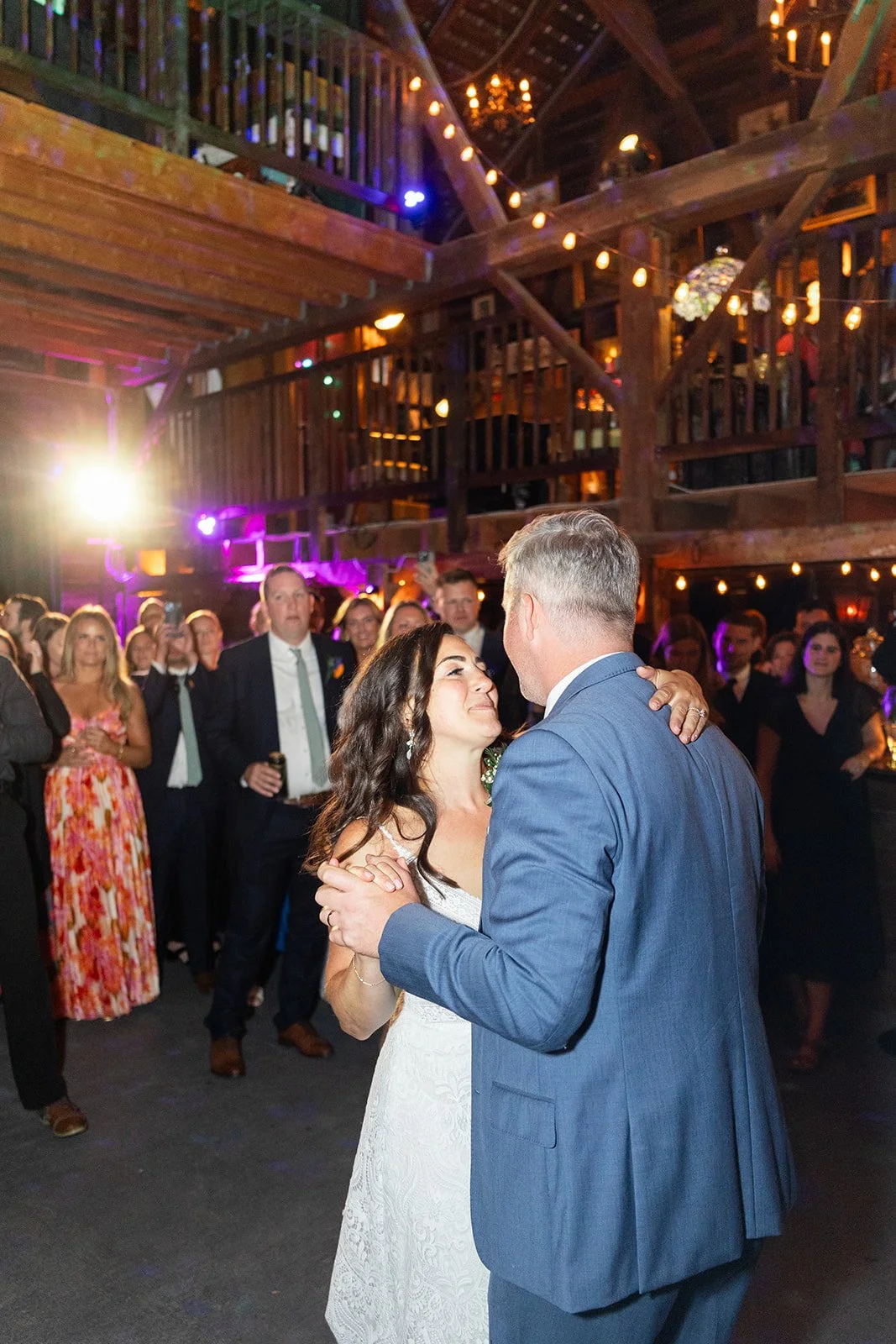 A bride and groom dance closely during their wedding reception in a rustic barn setting, surrounded by guests, with warm lighting and wooden decor.