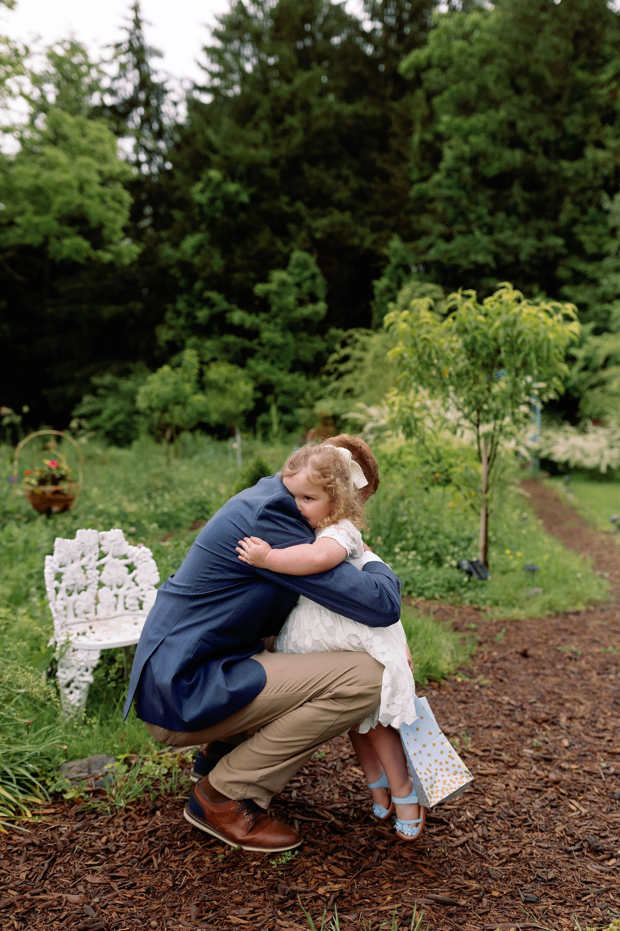 A man and young girl hugging each other in a garden, with the man crouching down and the girl holding a gift bag, surrounded by greenery and a small white decorative bench.