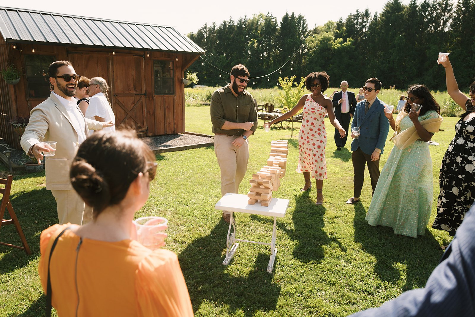 People outdoors playing a giant Jenga game with a wooden table and a rustic barn in the background, on a sunny day with green grass and trees.