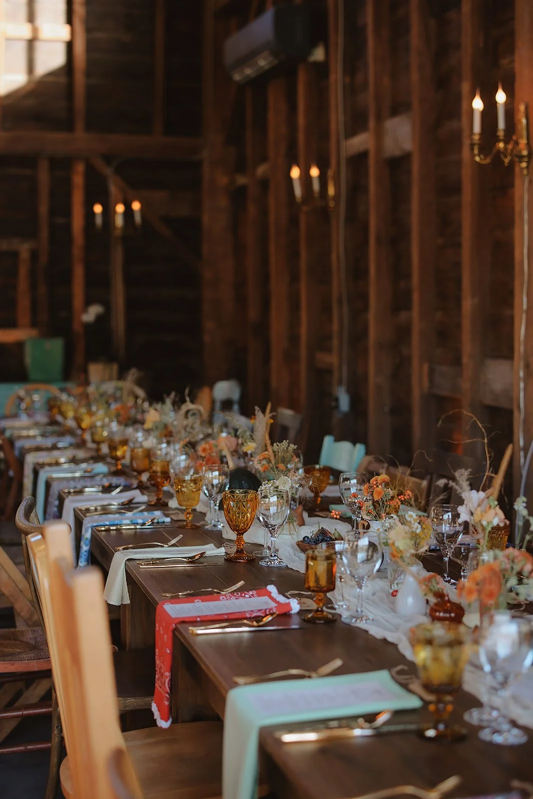 Long wooden banquet table decorated with floral centerpieces, various glassware, and set with plates and cutlery inside a rustic wooden barn.