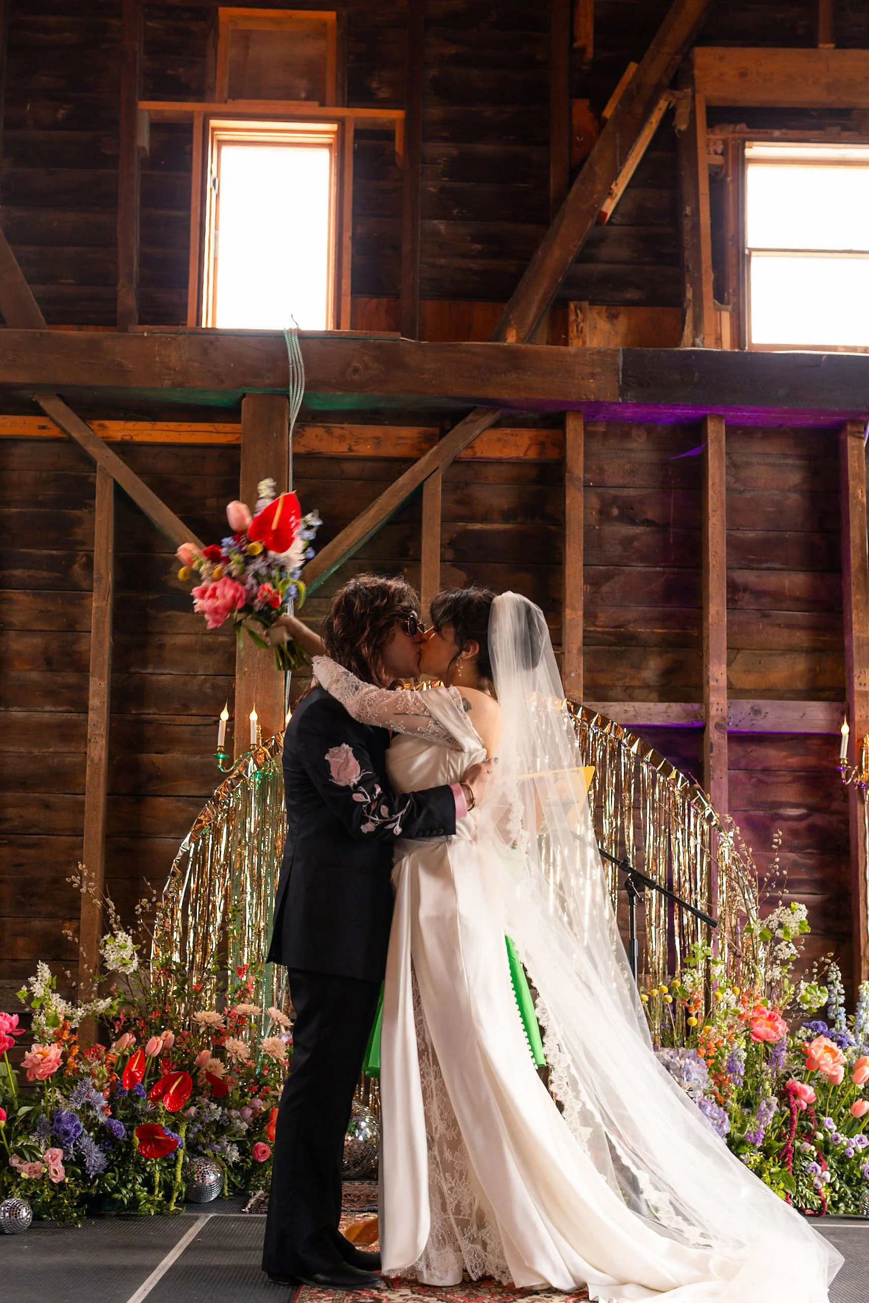 Couple kissing at wedding, holding bouquet, reception setup with flowers and candles, in rustic barn with wooden walls and loft.