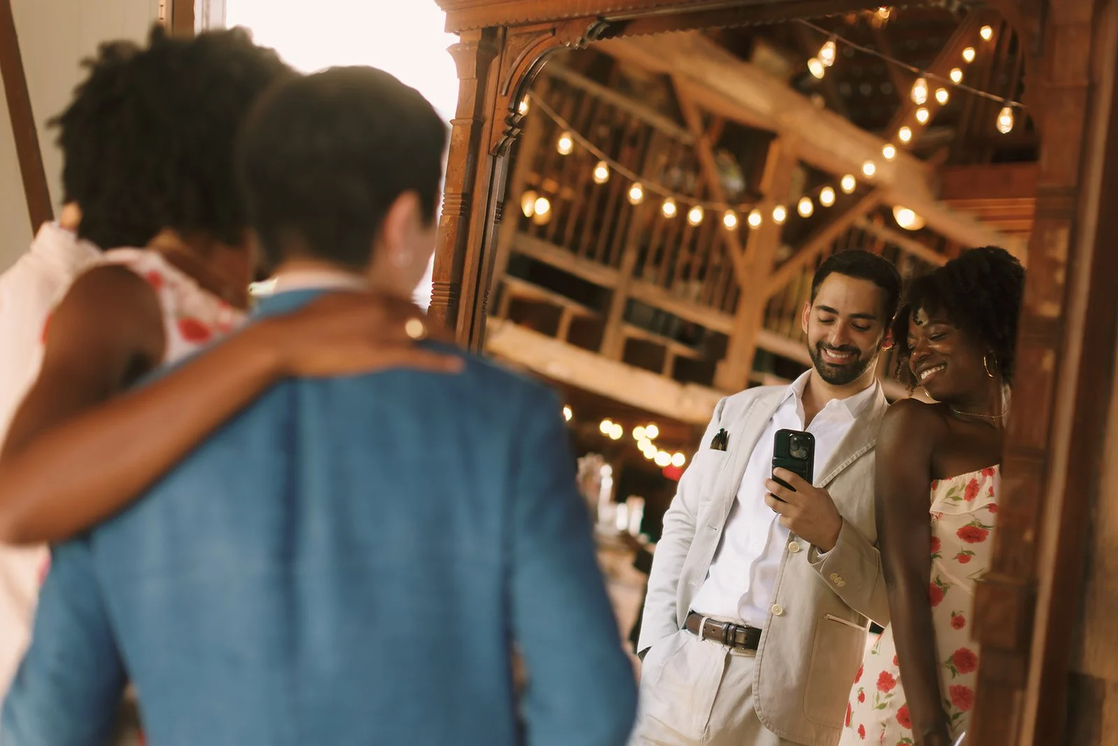 A group of four friends takes a selfie in front of a mirror at a rustic, warmly lit venue decorated with string lights, with smiles and friendly gestures.