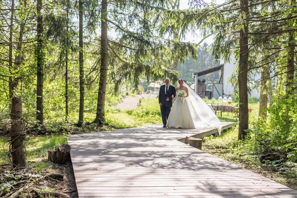 A bride and an older man walking on a wooden pathway in a forest, with a table set up for a wedding celebration in the background.