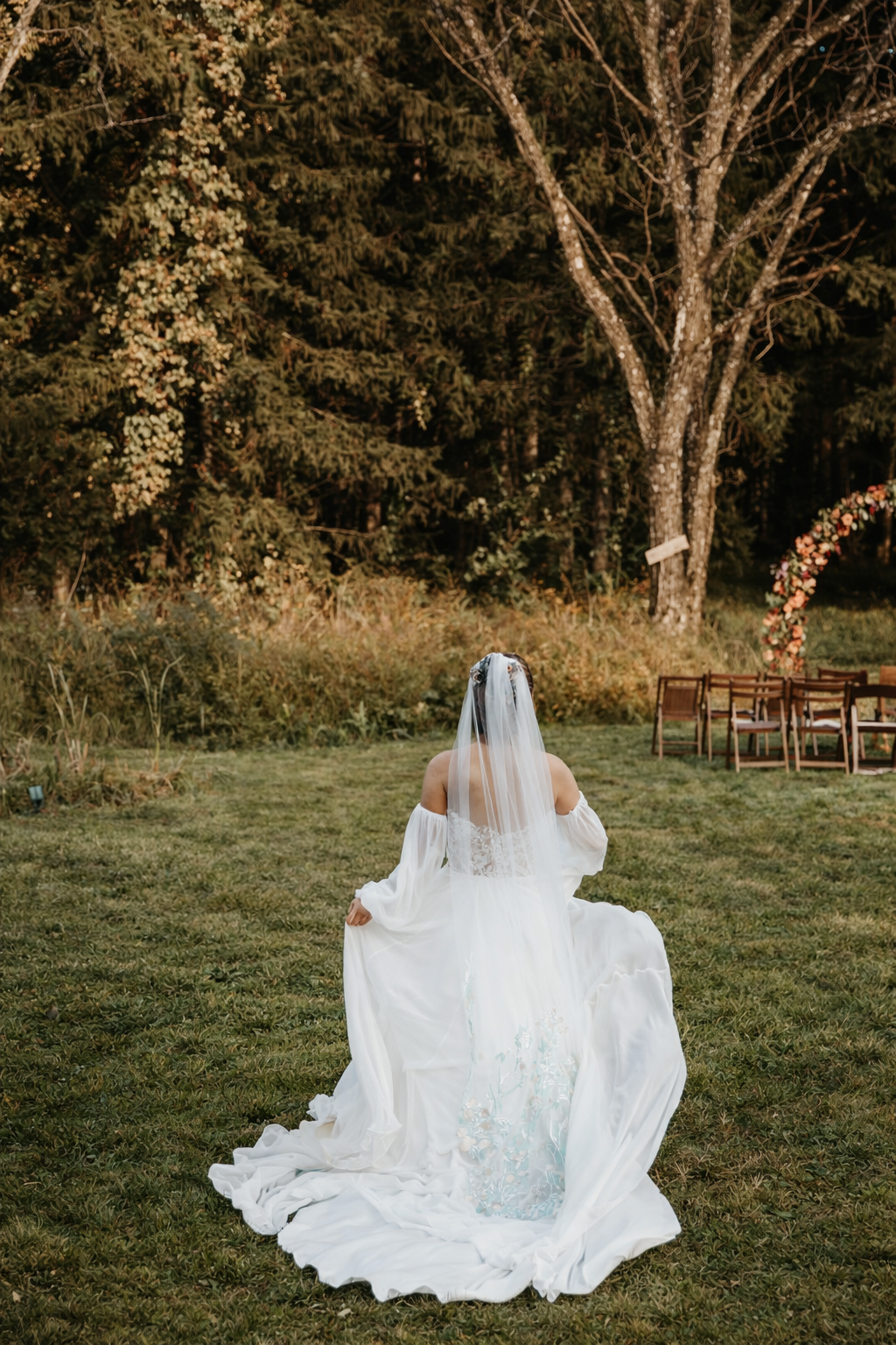 A bride in a white wedding dress and veil sits on the grass in an outdoor setting, with trees and chairs in the background.
