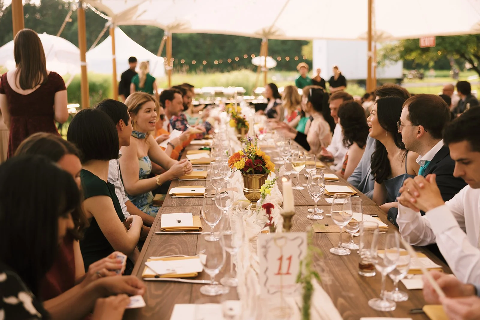 People seated at a long outdoor banquet table under a canopy during daytime, with floral centerpieces, wine glasses, and table settings visible.