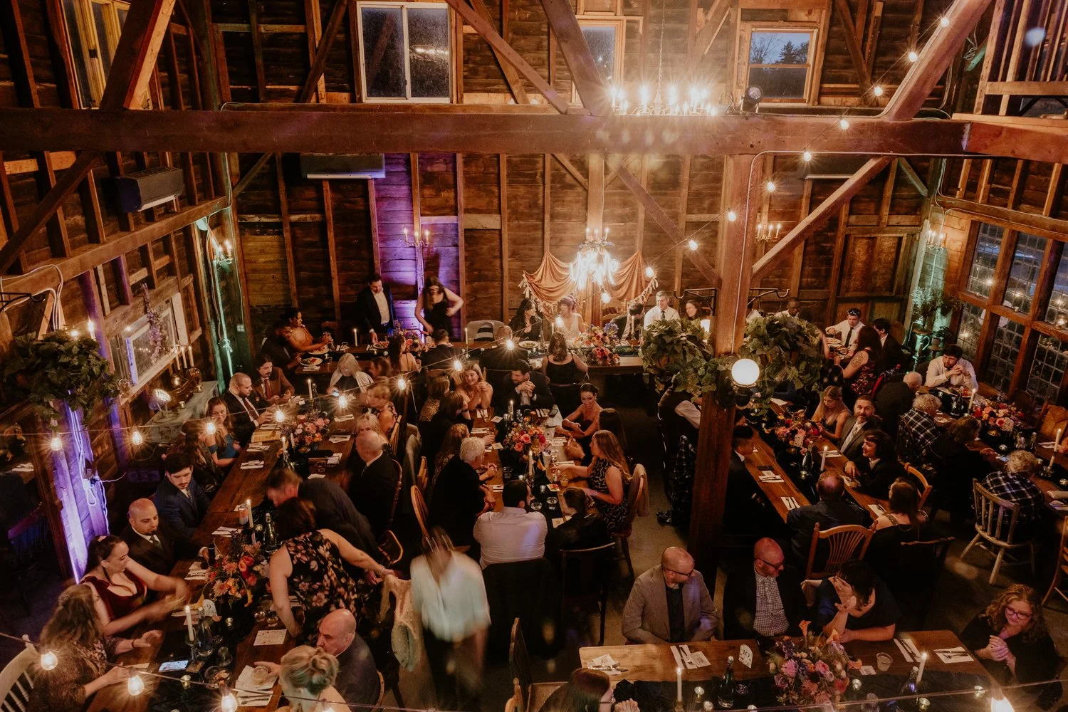 Guests seated at long tables decorated with flowers and candles in a rustic wooden barn, with a band or speakers at the front and chandeliers hanging from the ceiling, indicating a formal event or celebration.