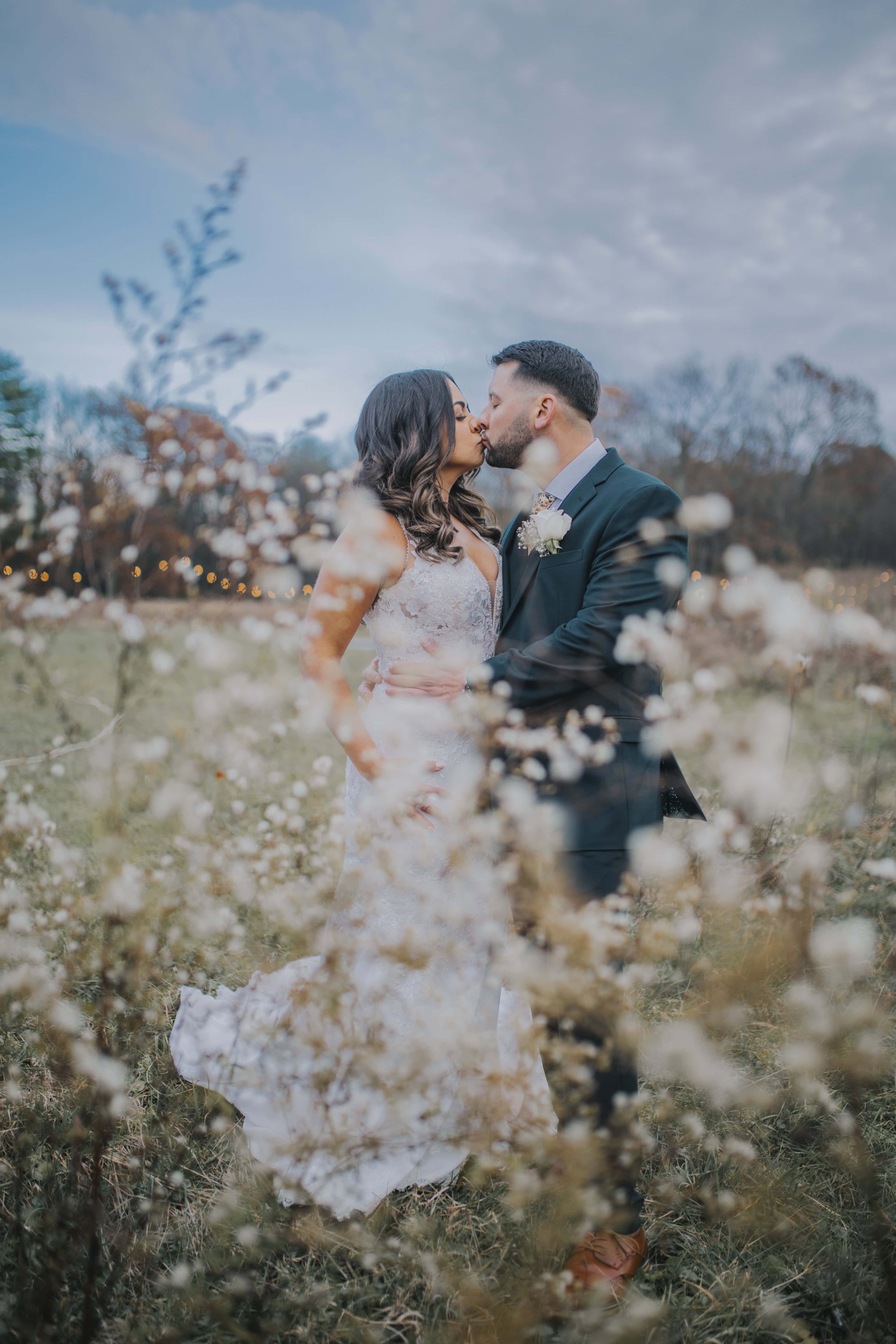 A newlywed couple sharing a kiss in a field of white flowers, with trees and fairy lights in the background.