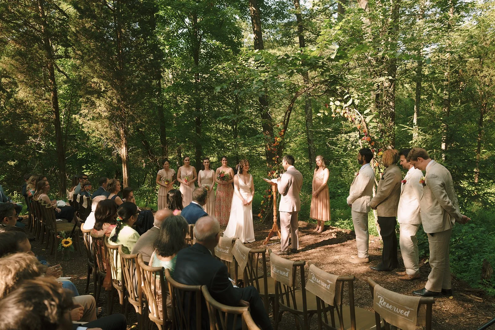 Outdoor wedding ceremony in a forest with officiant, bride, groom, bridesmaids, and groomsmen, surrounded by trees and green foliage.