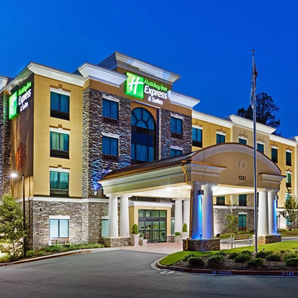 Exterior view of a hotel driveway  surrounded by bushes and parking spaces under a blue sky with clouds.