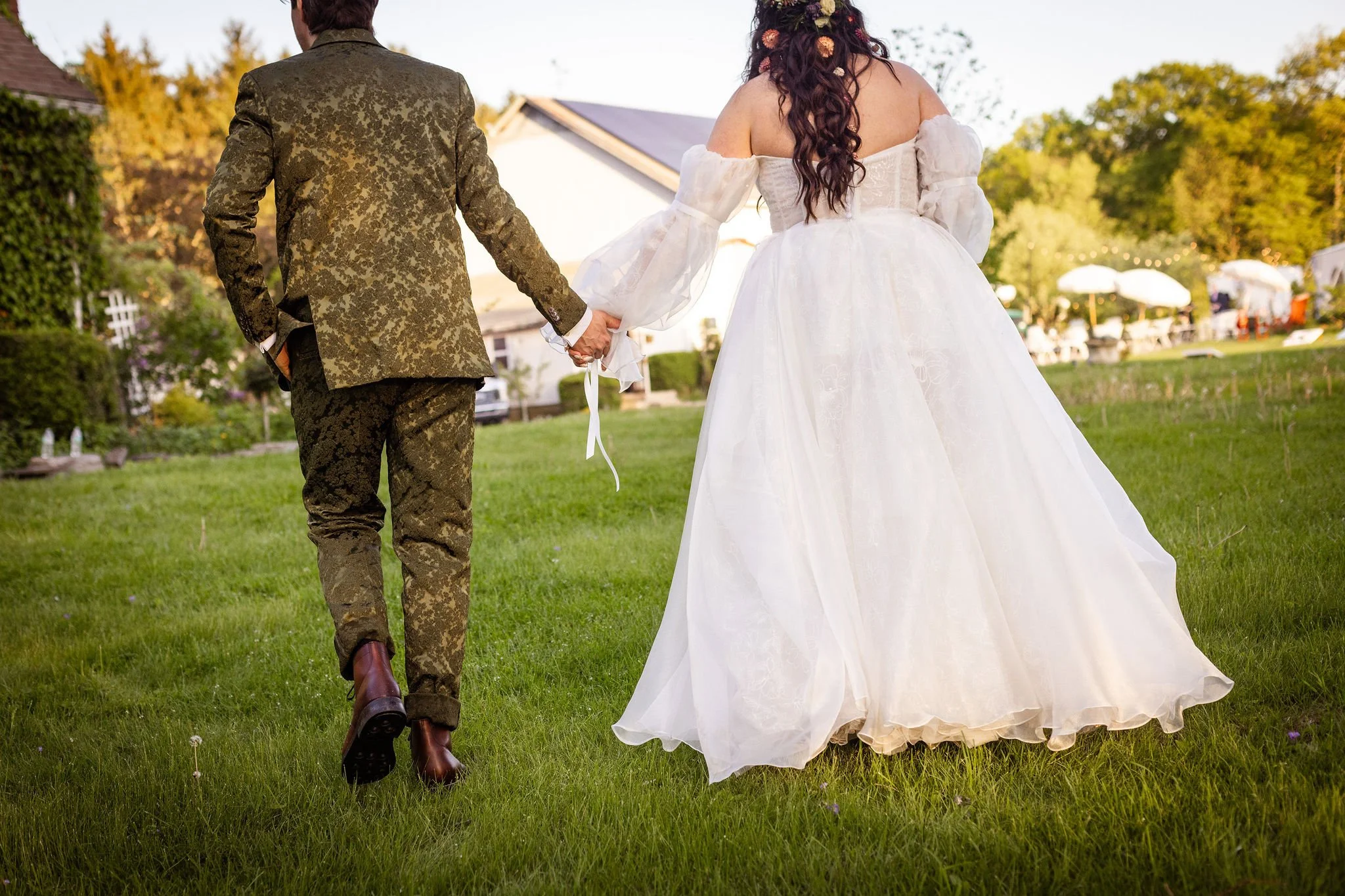 A bride and groom holding hands from behind, walking on grass outdoors during a wedding.