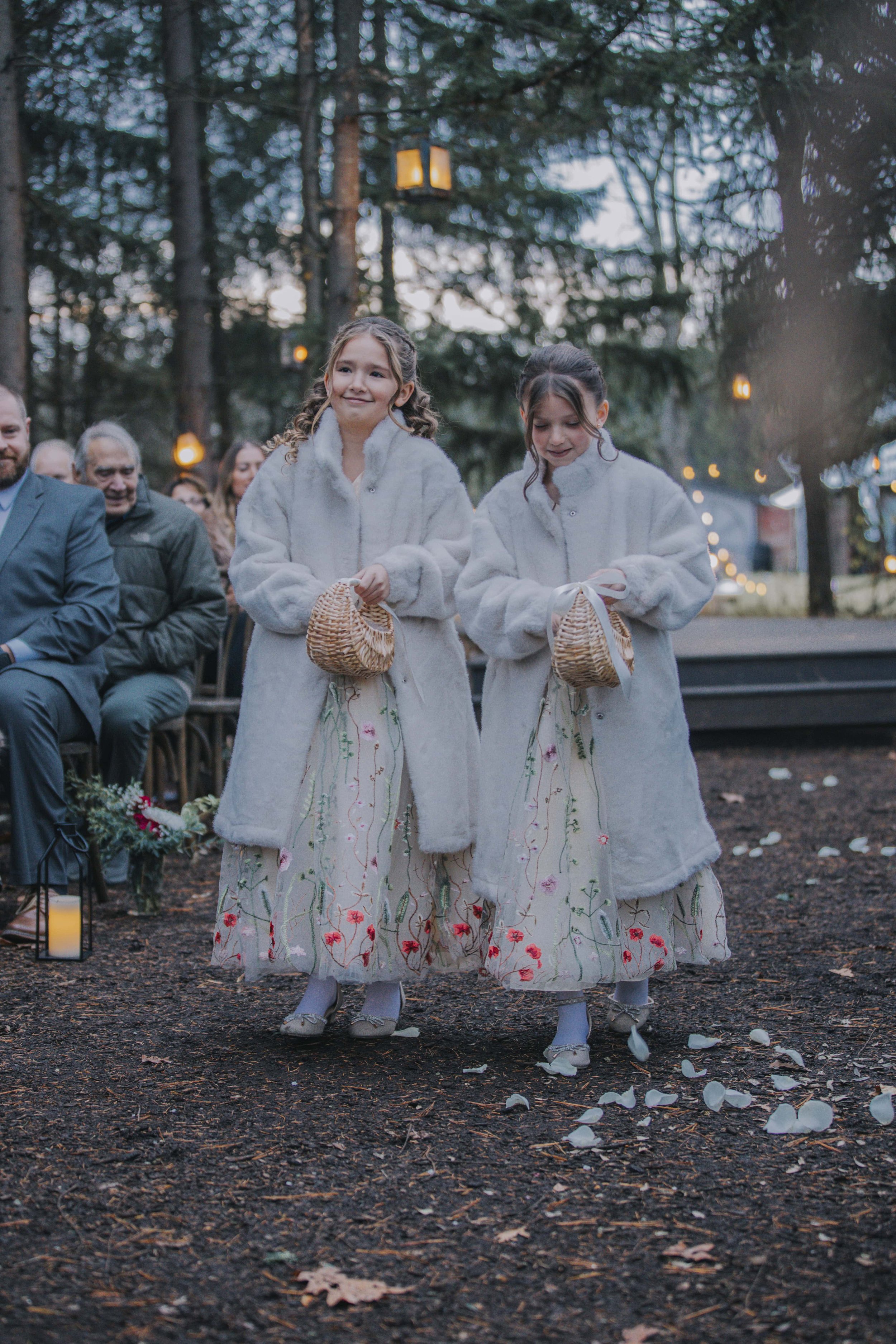 Two young girls in white fur coats and embroidered skirts holding small baskets walk on a dirt path at an outdoor event during the evening, with seated guests and string lights in the background.