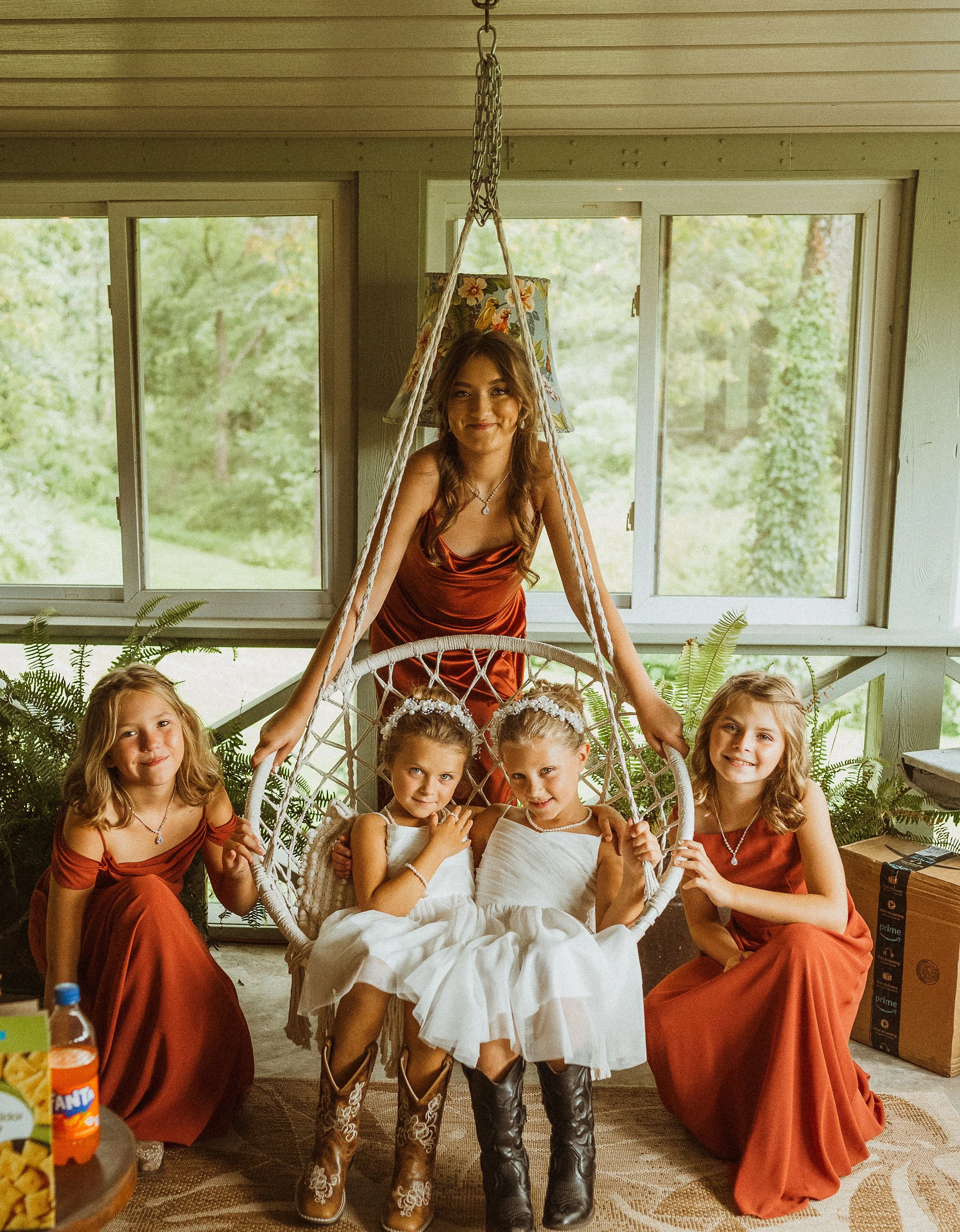A young woman with long, wavy brown hair wearing a satin red dress stands behind a white hanging chair, surrounded by four young girls. The girls wear white dresses with floral headbands, and two are in cowboy boots. They are inside a bright room wit