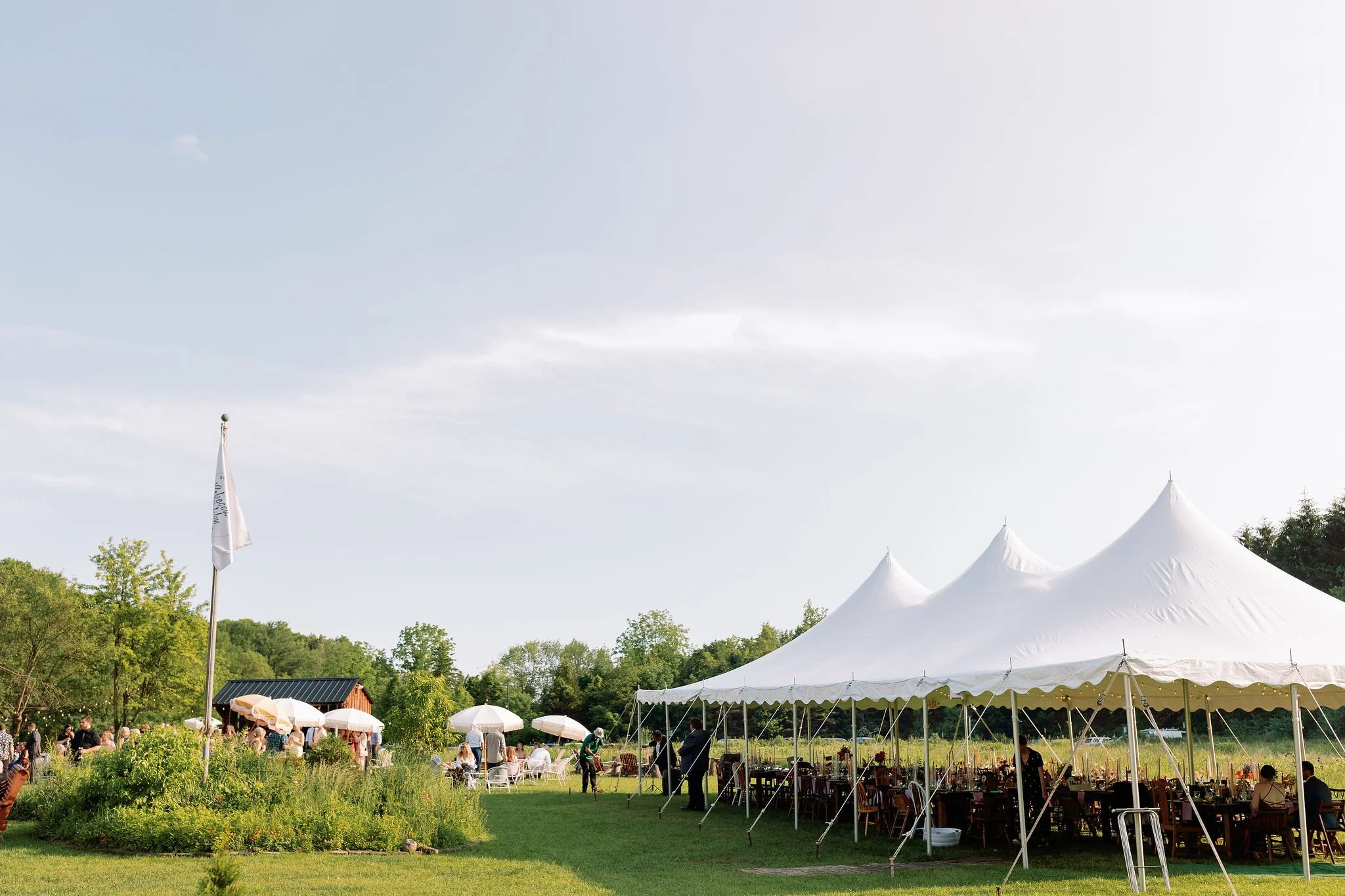 Outdoor event with white tents, tables, and people enjoying a sunny day on a grassy field.