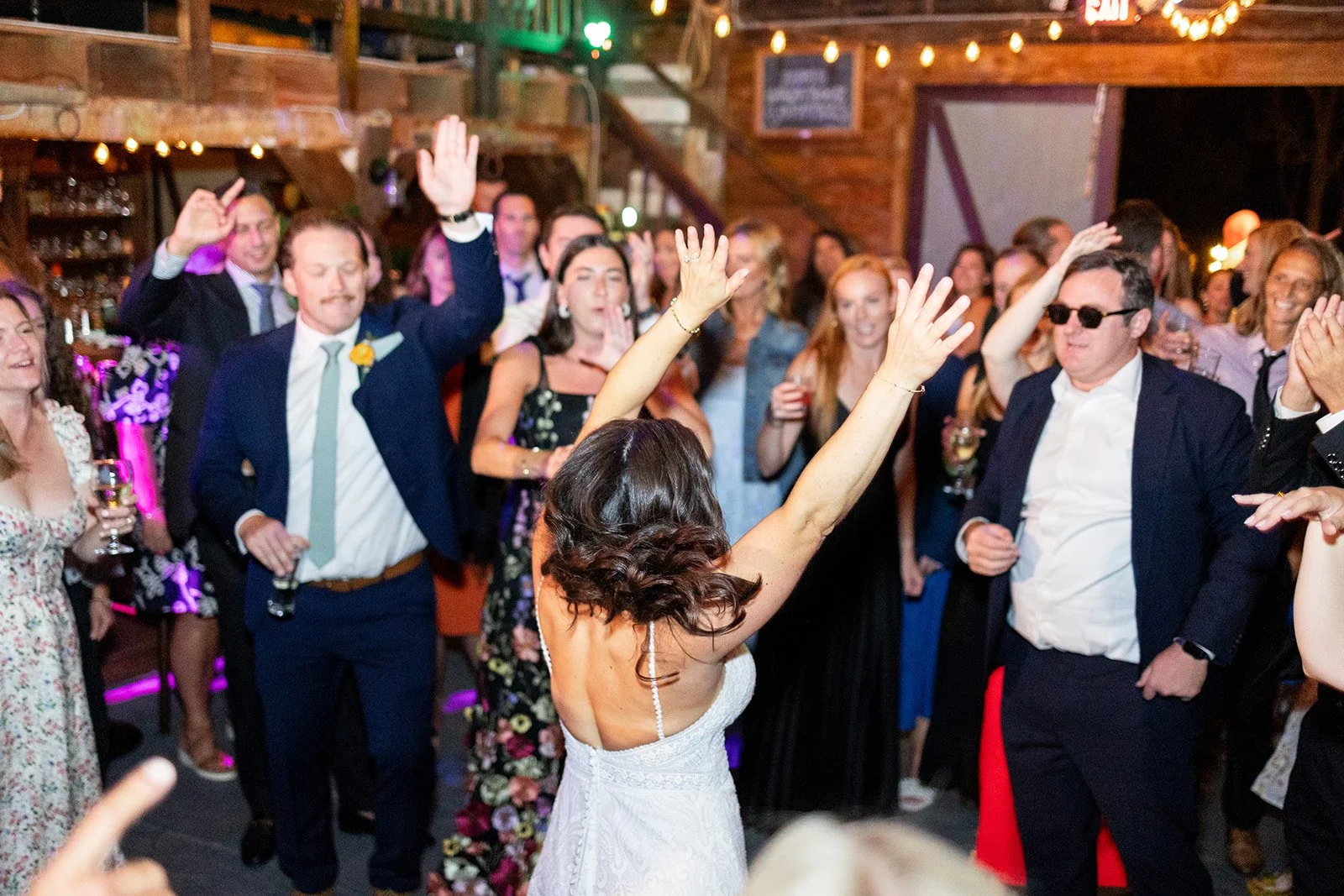 A bride dancing with her arms raised in front of a crowd at her wedding reception, with guests clapping and enjoying the music in a rustic indoor venue decorated with string lights.