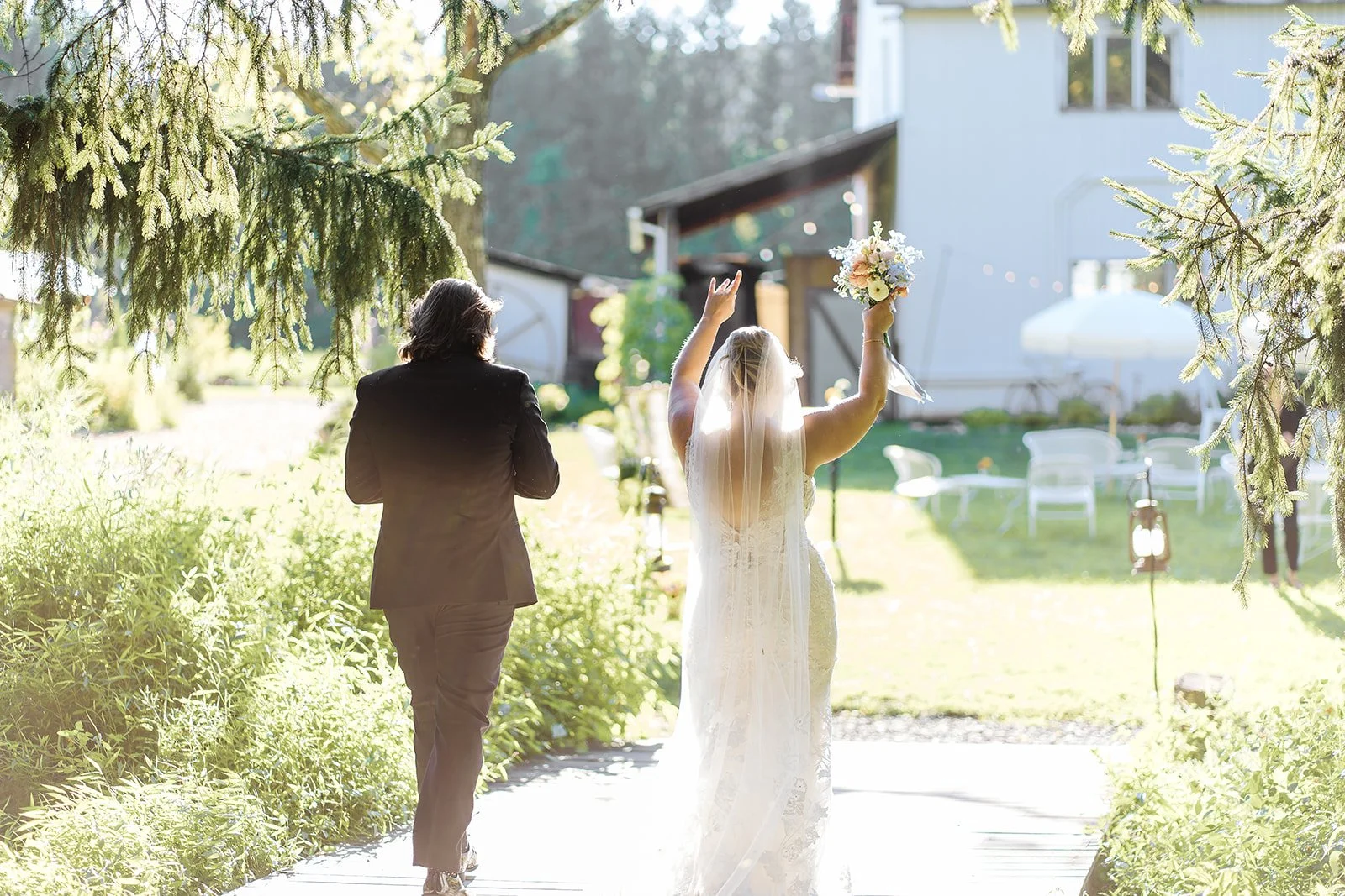 A bride in a white wedding dress and veil holding a bouquet of flowers with her arm raised, walking on a garden path with a man in a black suit, surrounded by greenery, trees, and outdoor wedding decorations.