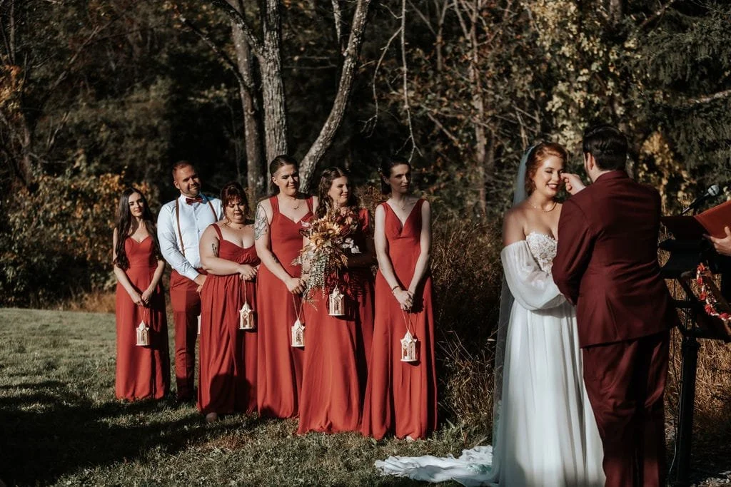 A wedding ceremony outdoors with six women in red dresses, a man and woman in wedding attire, and a photographer taking their picture in a wooded area.