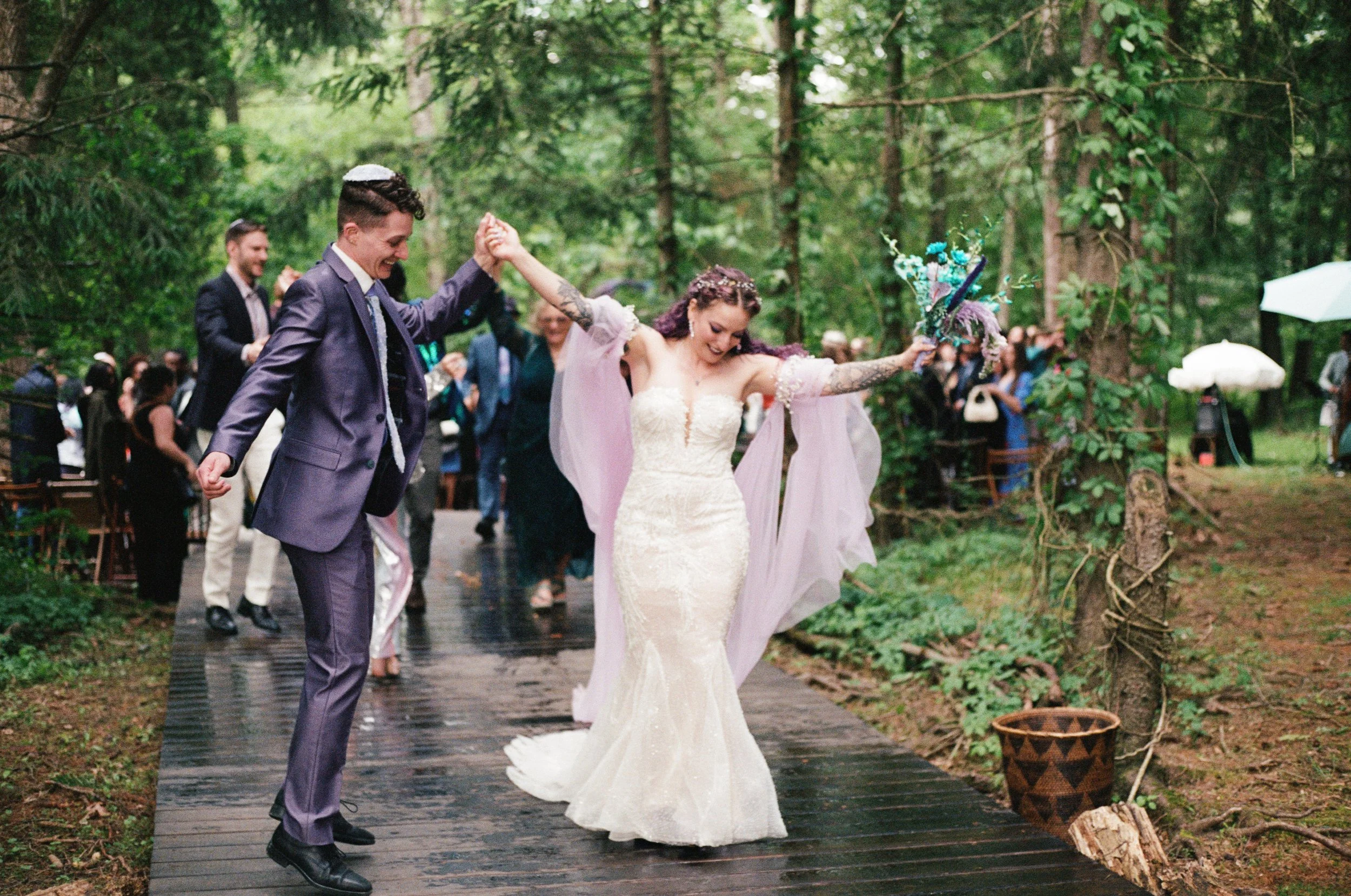 A bride and groom dancing outdoors on a wooden pathway during their wedding celebration surrounded by guests in a forest setting.