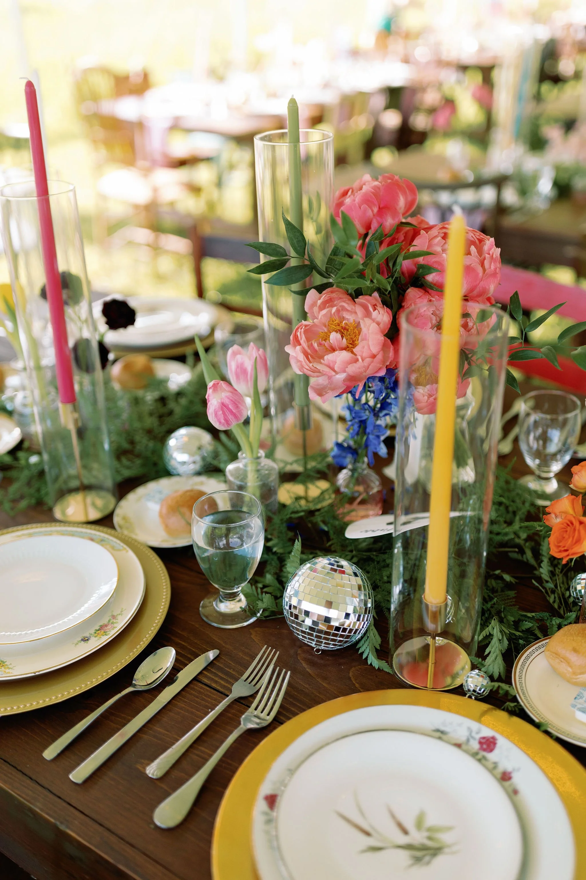 A decorated table setting with floral arrangements, tall colored candles in glass holders, and dinnerware including patterned plates, forks, knives, and glasses, set for a celebration or gathering.