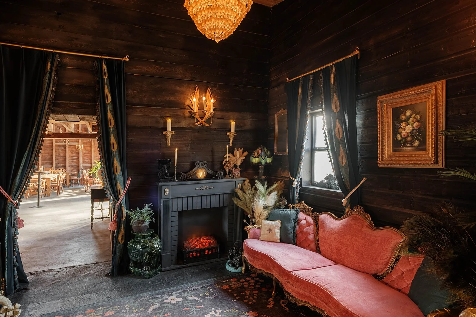 Cozy vintage living room with dark wood-paneled walls, pink velvet tufted sofa, and black fireplace, decorated with candles, plants, and art, illuminated by chandelier and wall sconce lights, with curtains, window, and framed floral art.