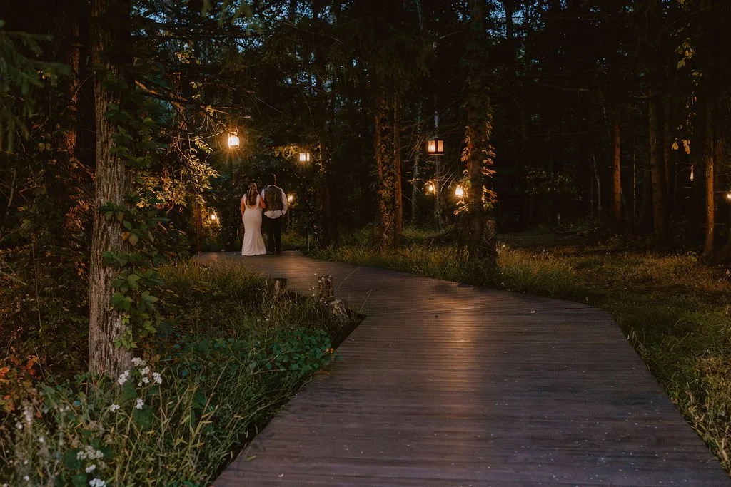 A couple dressed in wedding attire walking down a wooden path through a wooded area decorated with hanging lanterns at night.