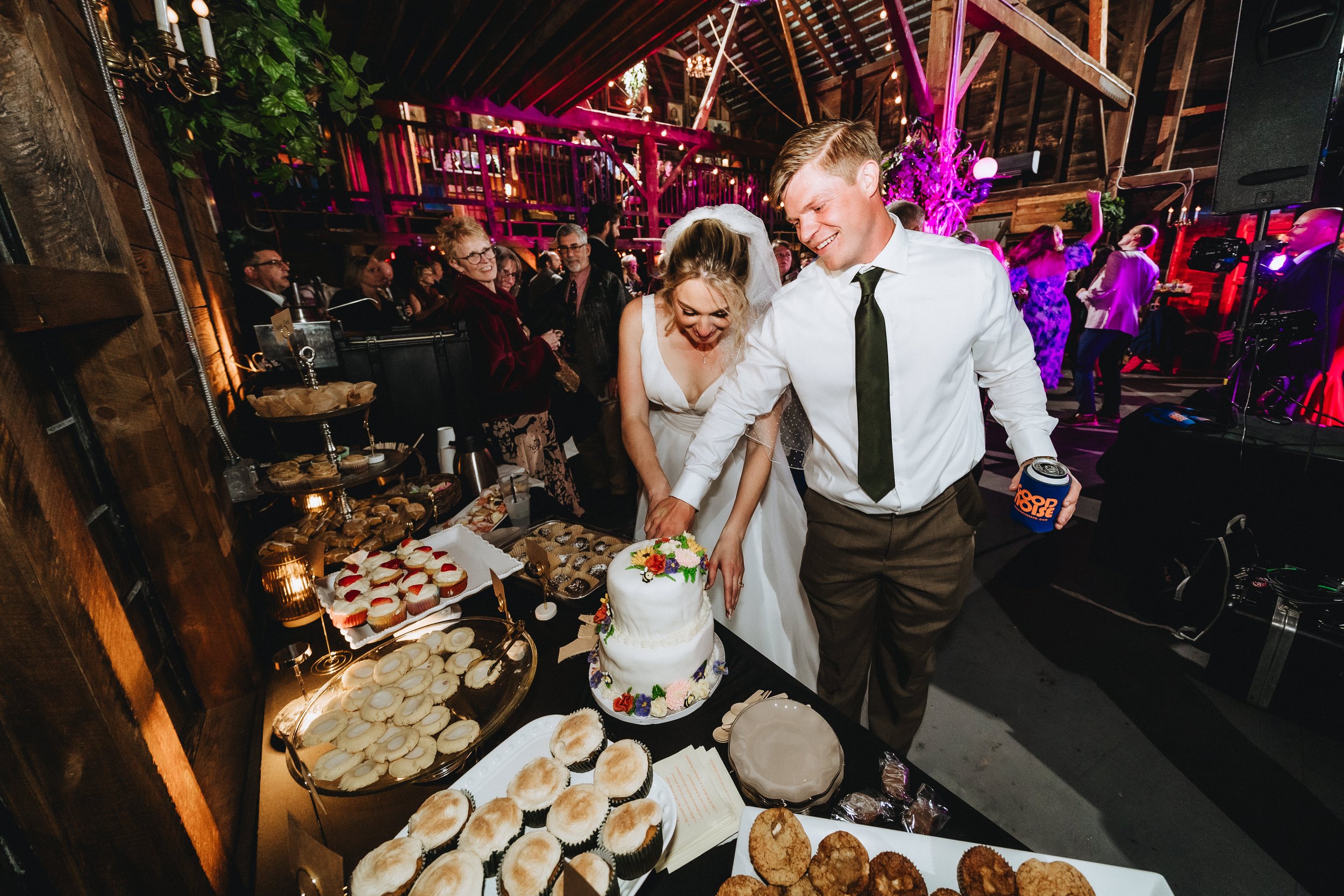 A bride and groom cutting their wedding cake at a reception with guests around them, a table full of desserts in the foreground, and a lively dance floor with purple and pink lighting in the background.