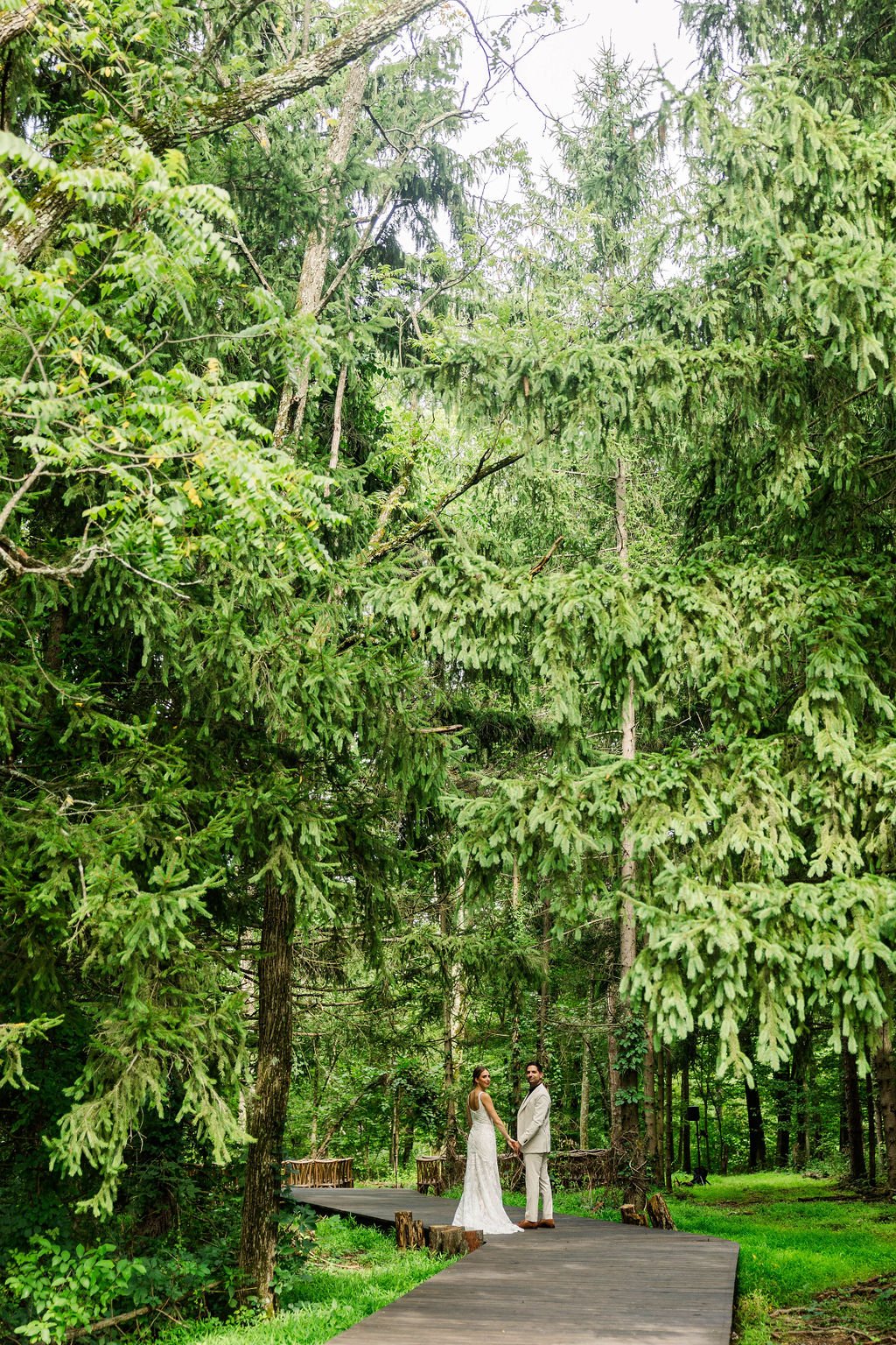 A couple in wedding attire holding hands, standing on a wooden path in a dense, green forest.