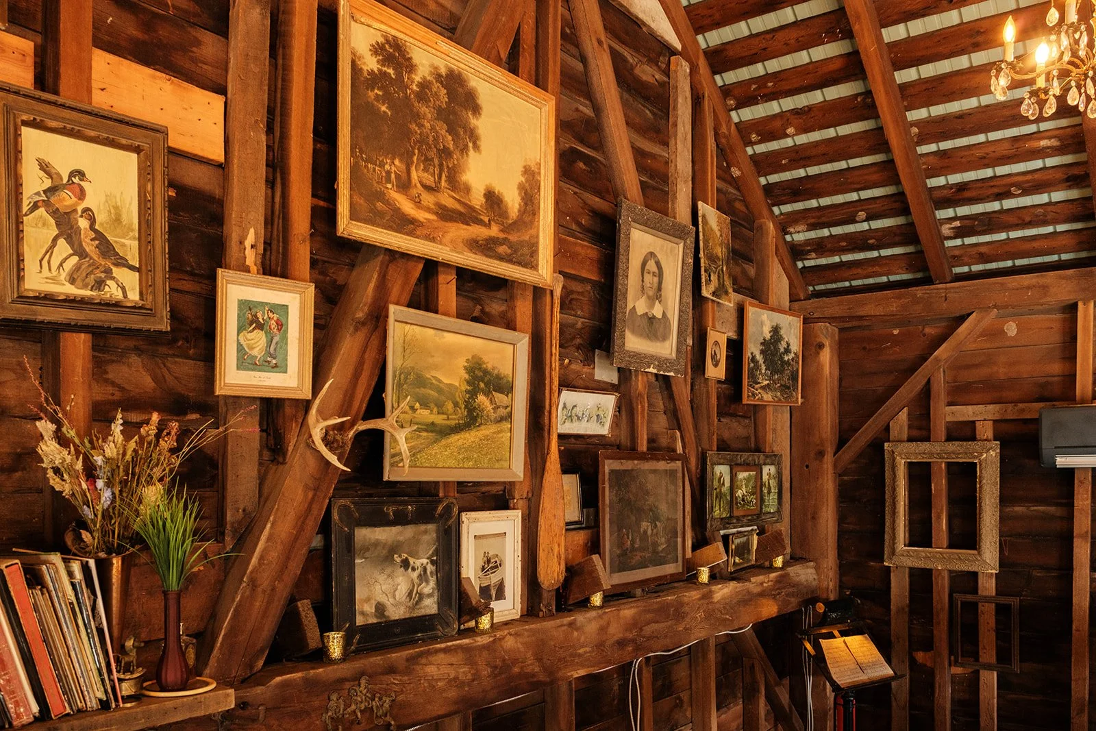Interior view of a rustic wooden wall decorated with various framed paintings and photographs, a bouquet of dried flowers in a vase, a stack of books, and a deer antler. There is a chandelier hanging from the ceiling.