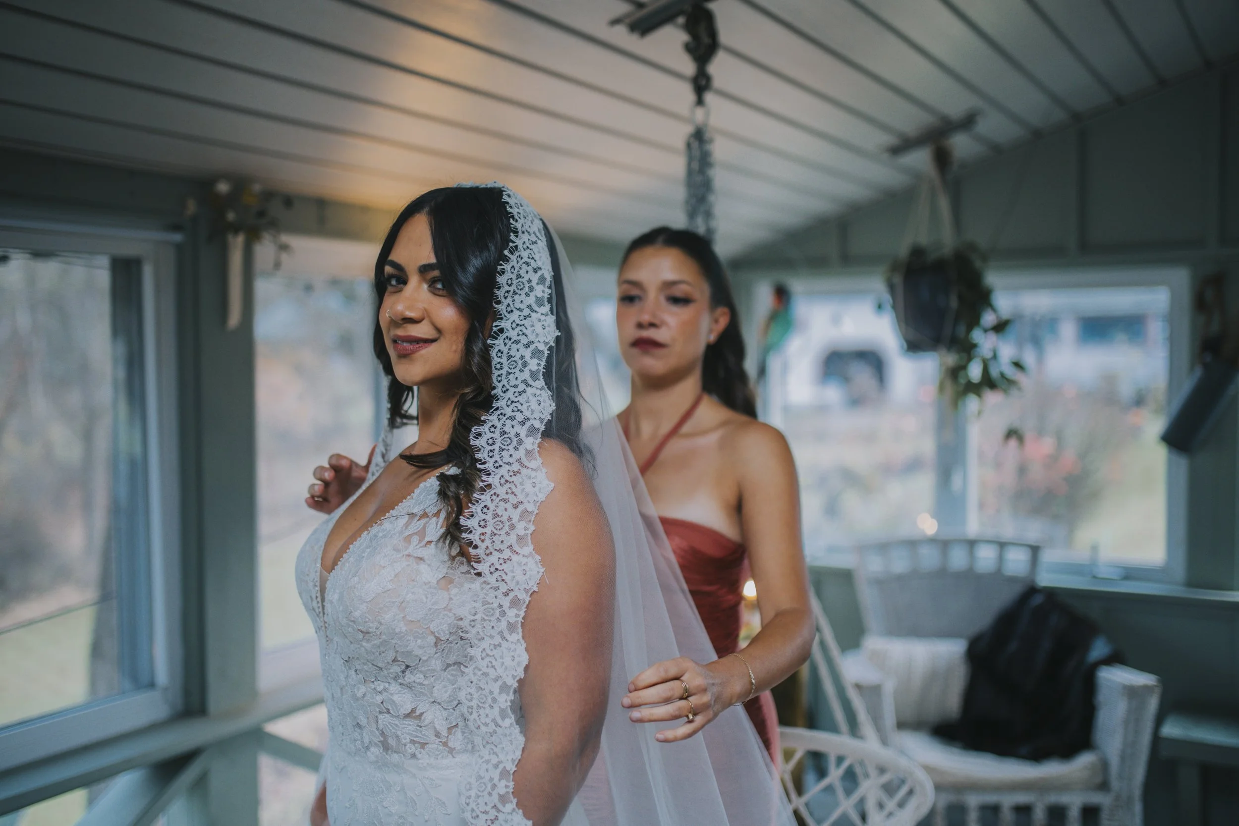 A bride in a white lace wedding dress with a lace veil is being assisted by a woman in a strapless red dress inside a sunlit room with large windows, outdoor scenery visible through the windows, and wicker furniture.
