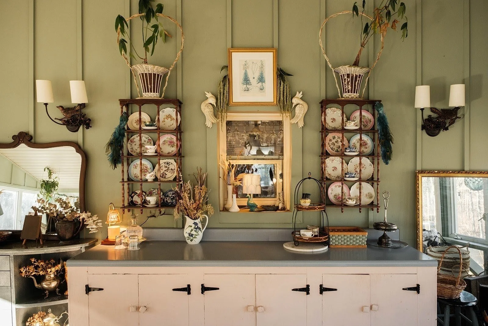 Decorative interior with green paneled walls, displaying shelves with teacups and plates, a vase with flowers, framed artwork, mirror, and vintage lighting fixtures.