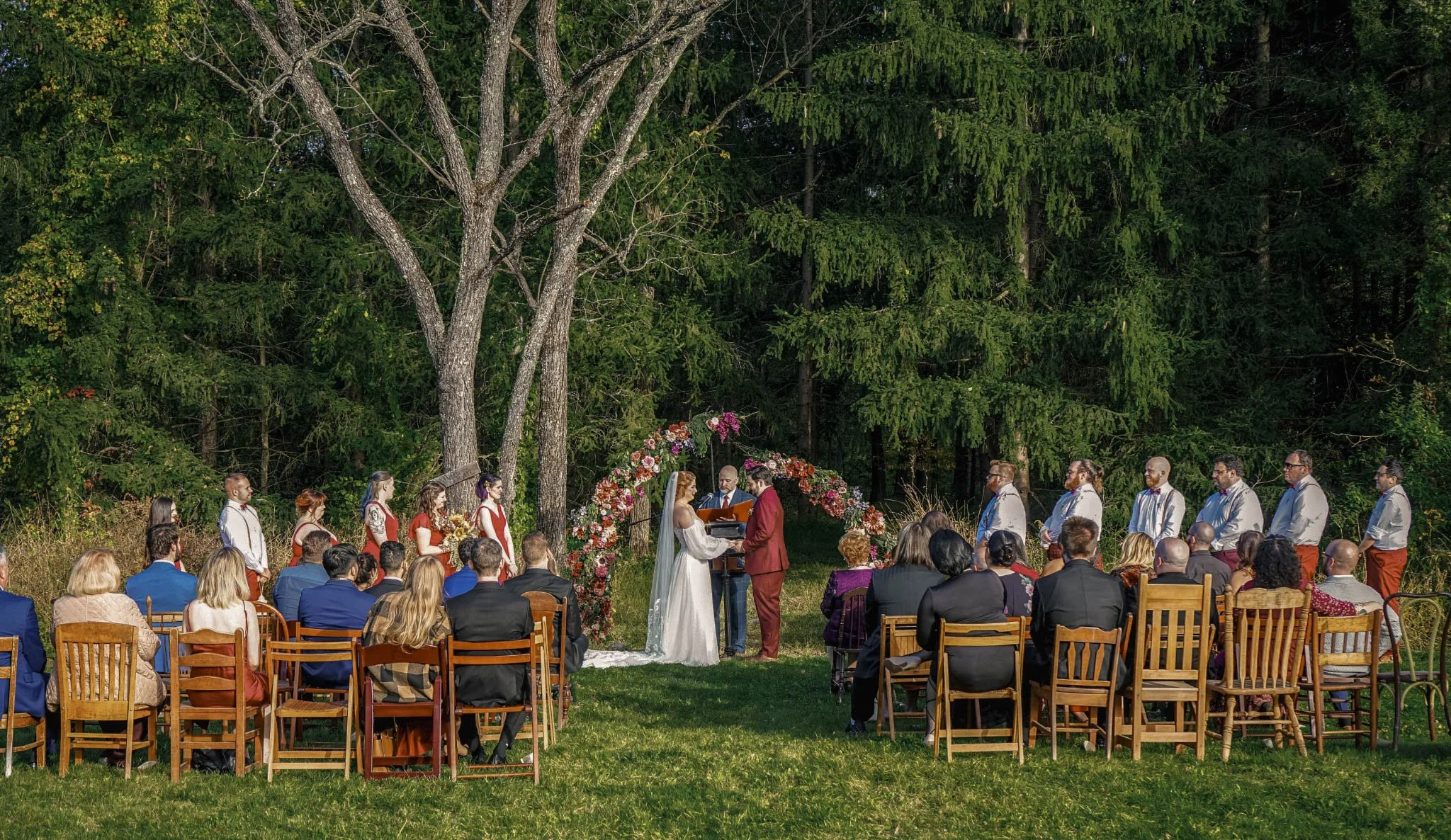 A wedding ceremony taking place outdoors in a green wooded area with a flower arch. The bride and groom are at the altar, with guests seated on wooden chairs facing them.