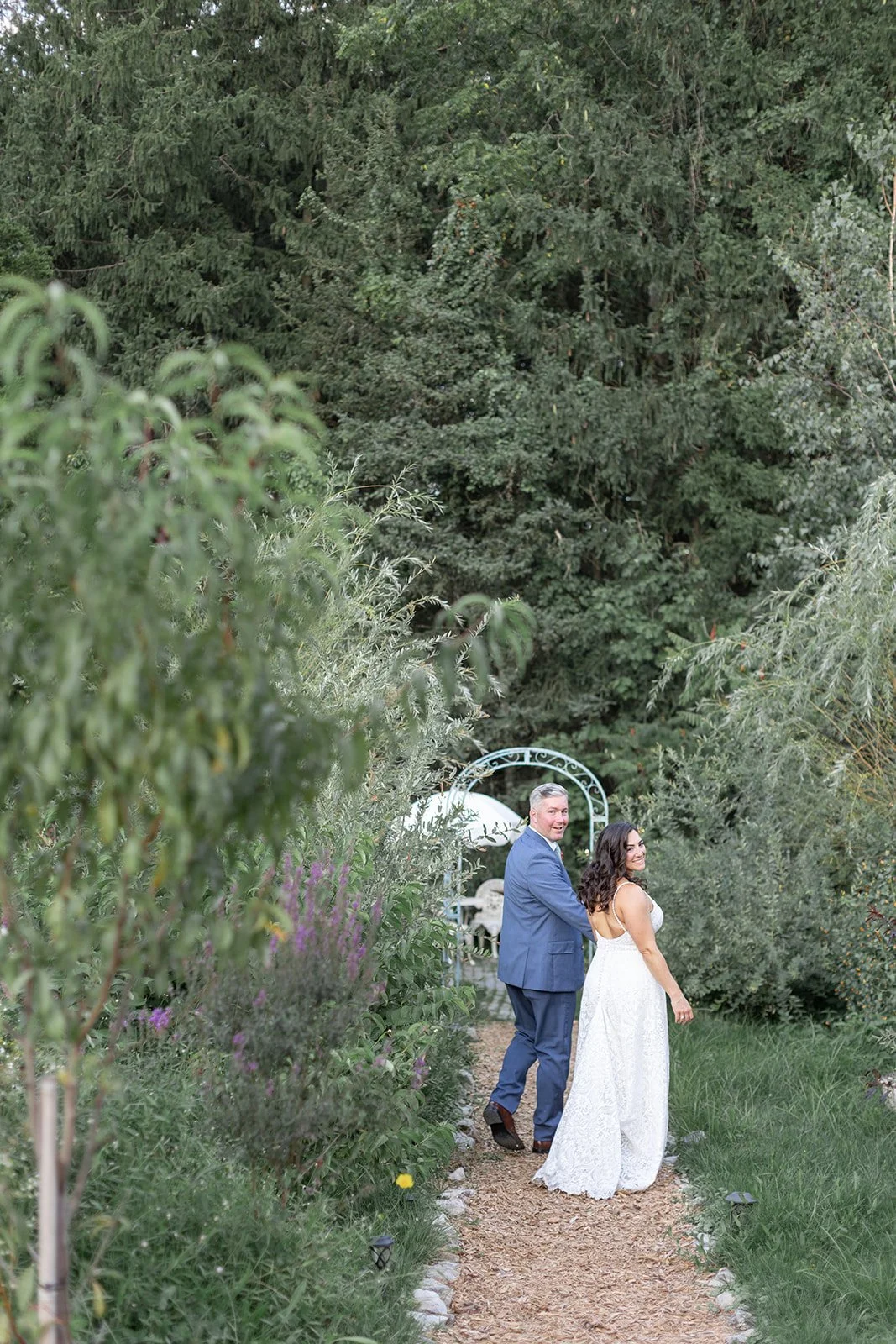 A couple, dressed in wedding attire, holding hands and walking on a garden pathway surrounded by lush greenery and plants, with a decorative arch in the background.