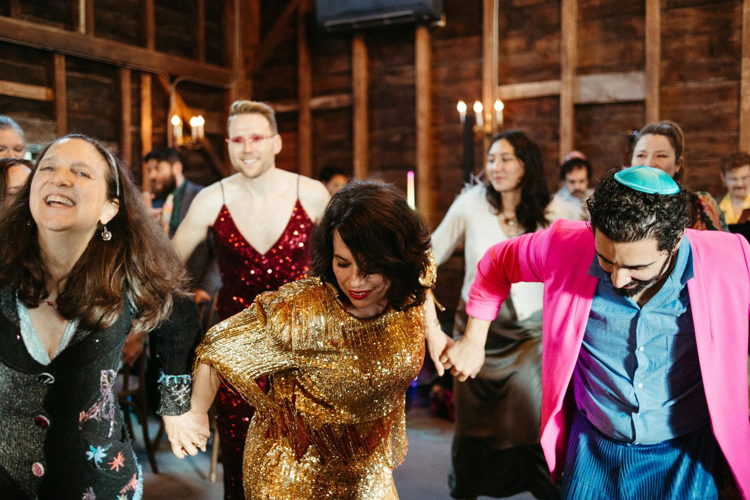 People dancing together at a celebration, with some wearing colorful, shiny, and festive clothing in a warmly lit wooden interior.