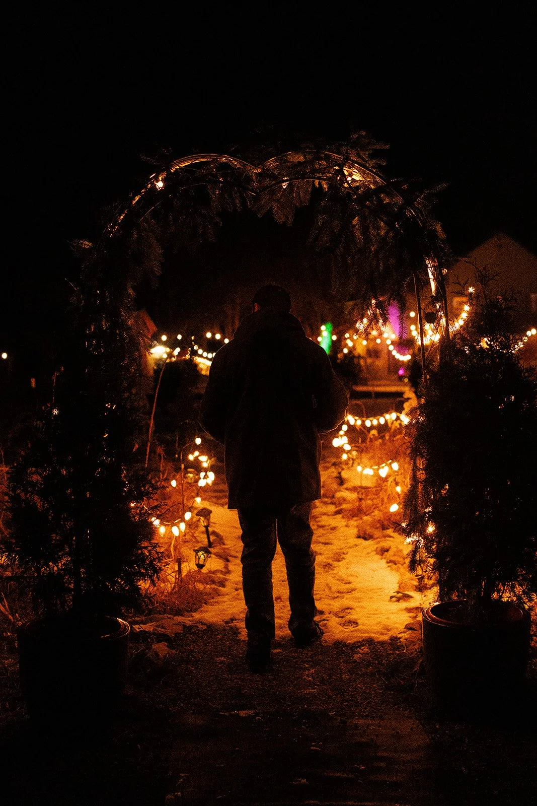 Person standing on a snow-covered pathway at night, illuminated by string lights and festive decorations.