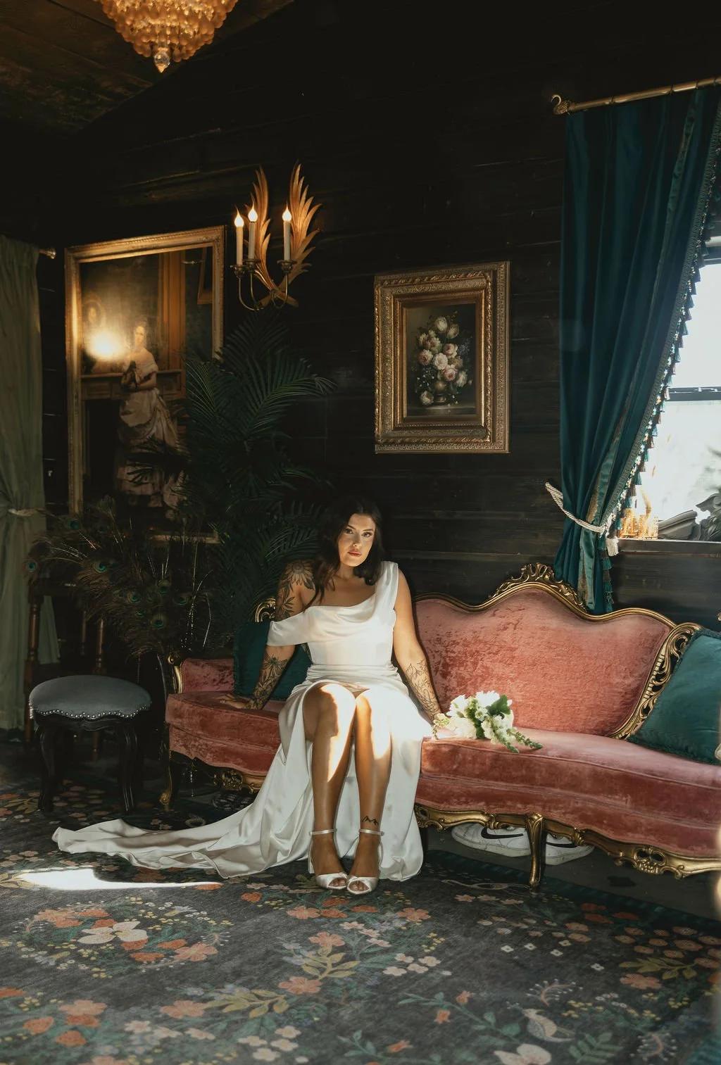Woman in white dress sitting on a vintage pink velvet sofa in an elegant room with dark wooden walls, framed paintings, and heavy teal curtains, holding a bouquet of white flowers.