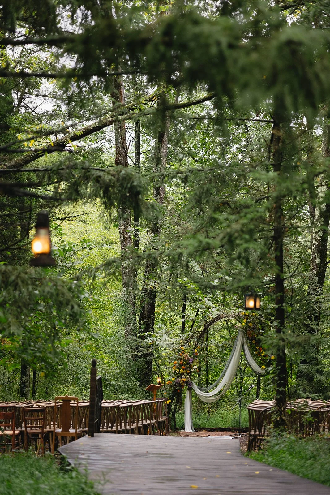 Outdoor wedding ceremony area set in a forest with chairs lining a wooden aisle, floral arch, draped fabric, lanterns hanging from trees, and a natural, green surroundings.