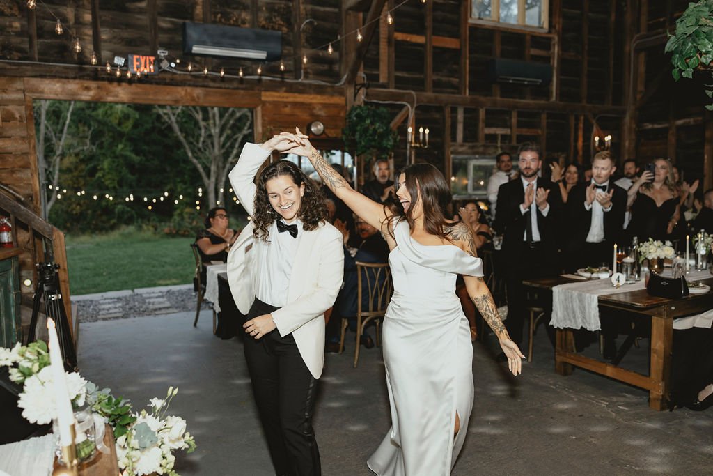 Two women dancing at a wedding reception, one in a tuxedo jacket and the other in a white dress, with guests clapping in the background inside a rustic wooden venue.