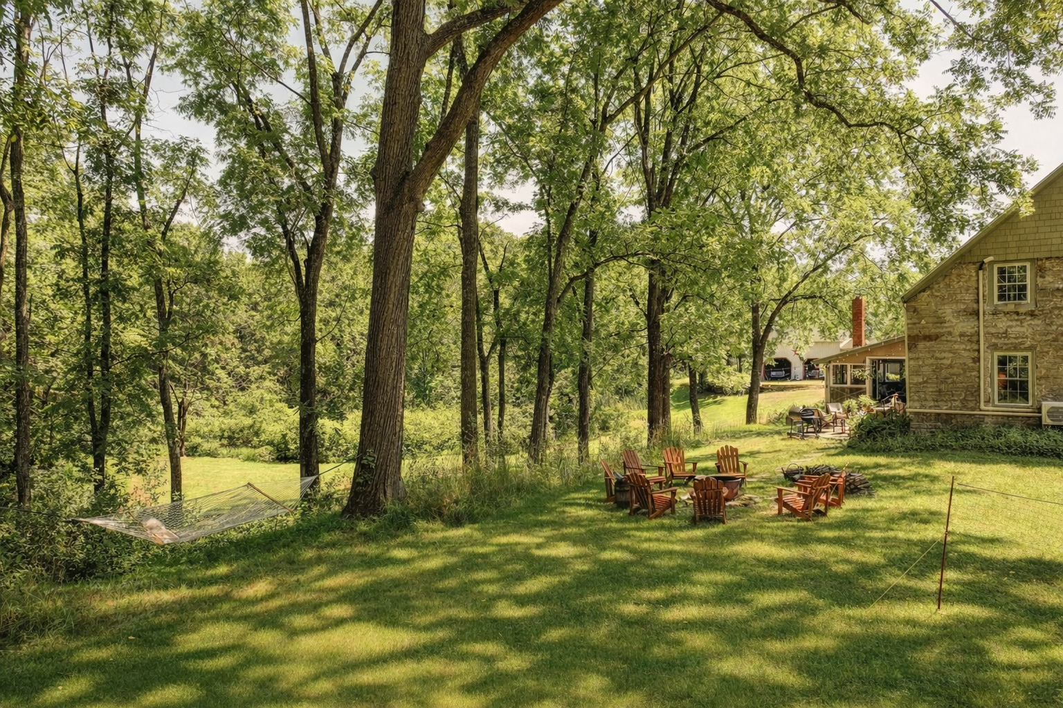 A backyard with green grass, several tall trees, an outdoor seating area with wooden chairs, and a house with a stone exterior.