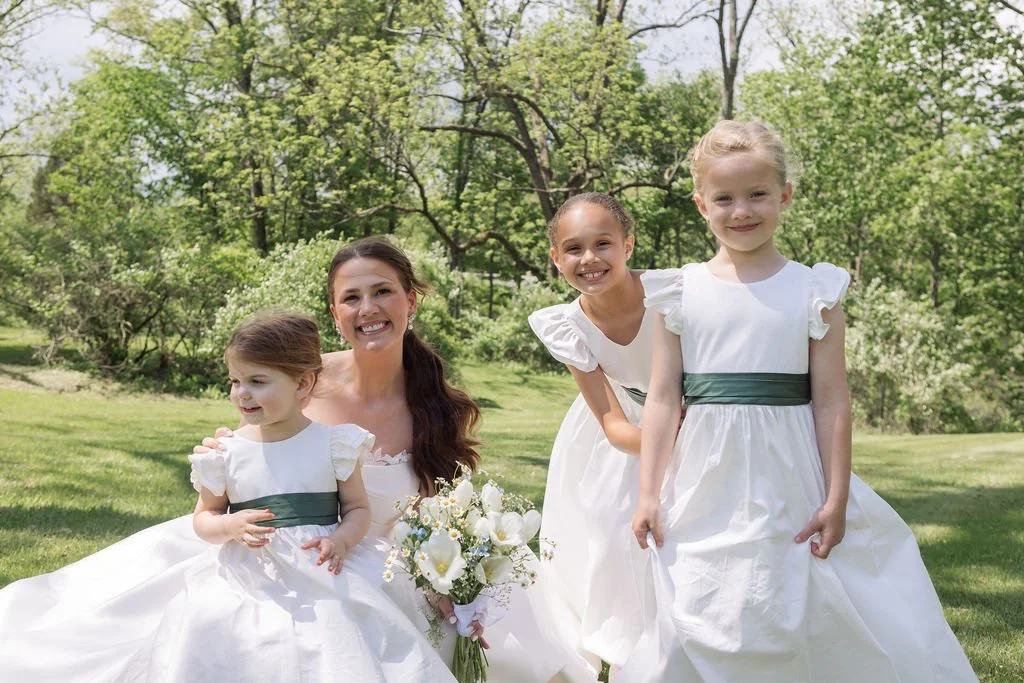 Four females, one adult woman and three young girls, dressed in white dresses with dark green sashes, sitting and standing on grass in a park or garden with green trees in the background, smiling.