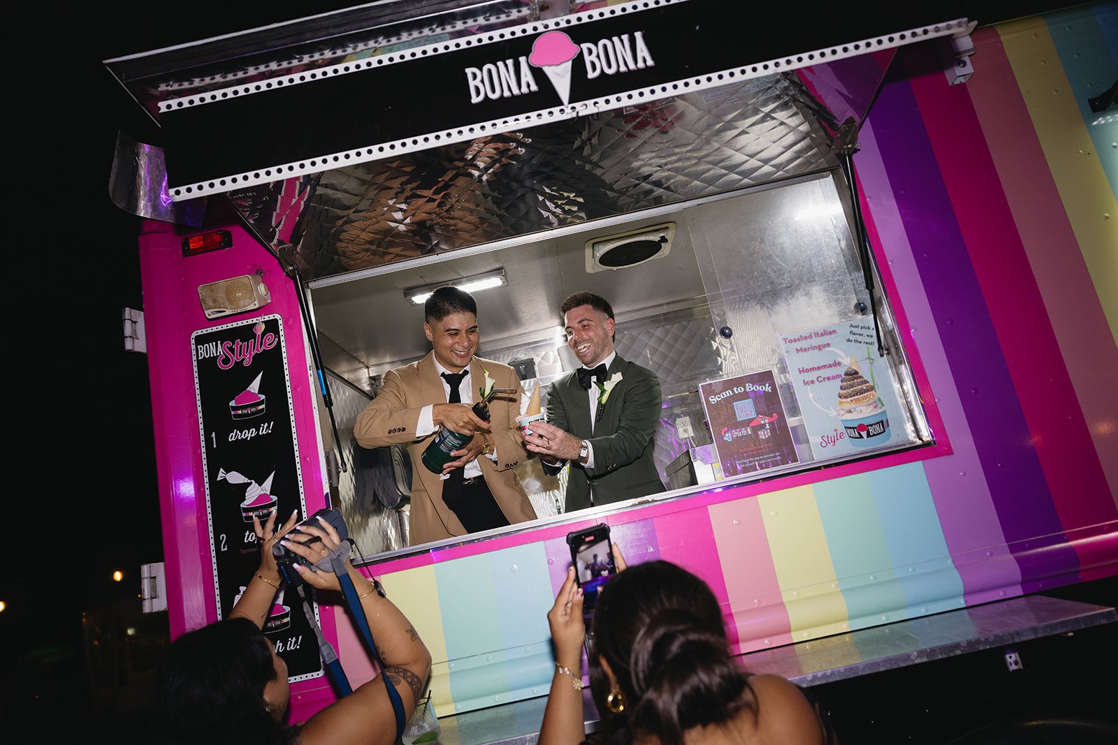 Two men in tuxedos celebrating inside a colorful pink ice cream truck called "BONA BONA" at night, with one pouring a drink into a cone held by the other. Several people outside are taking pictures of the moment.