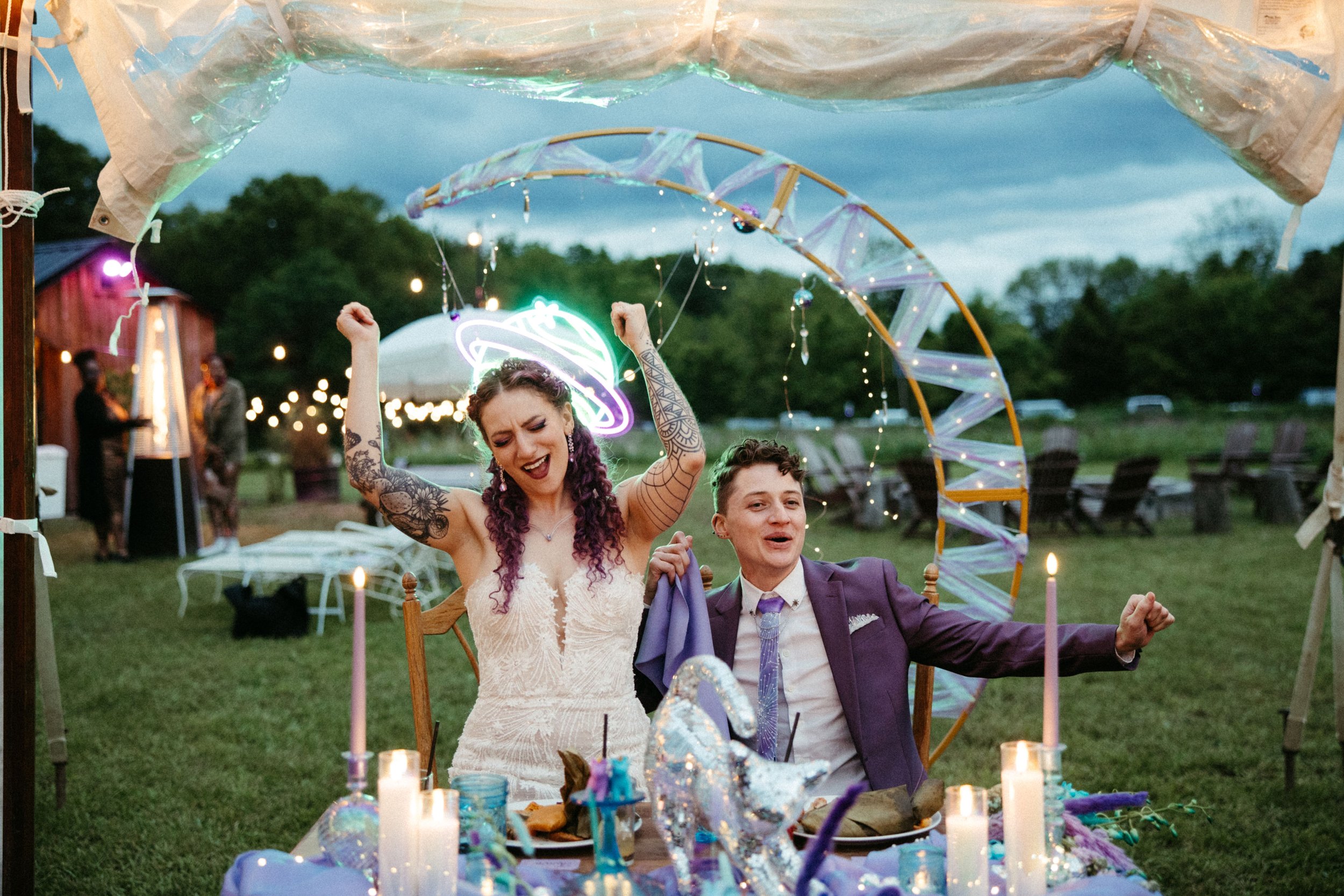 Two people celebrating at an outdoor party with fairy lights and candles on a decorated table, one woman with tattoos raising her arms in celebration and a man in a purple suit sitting beside her, both appearing happy.